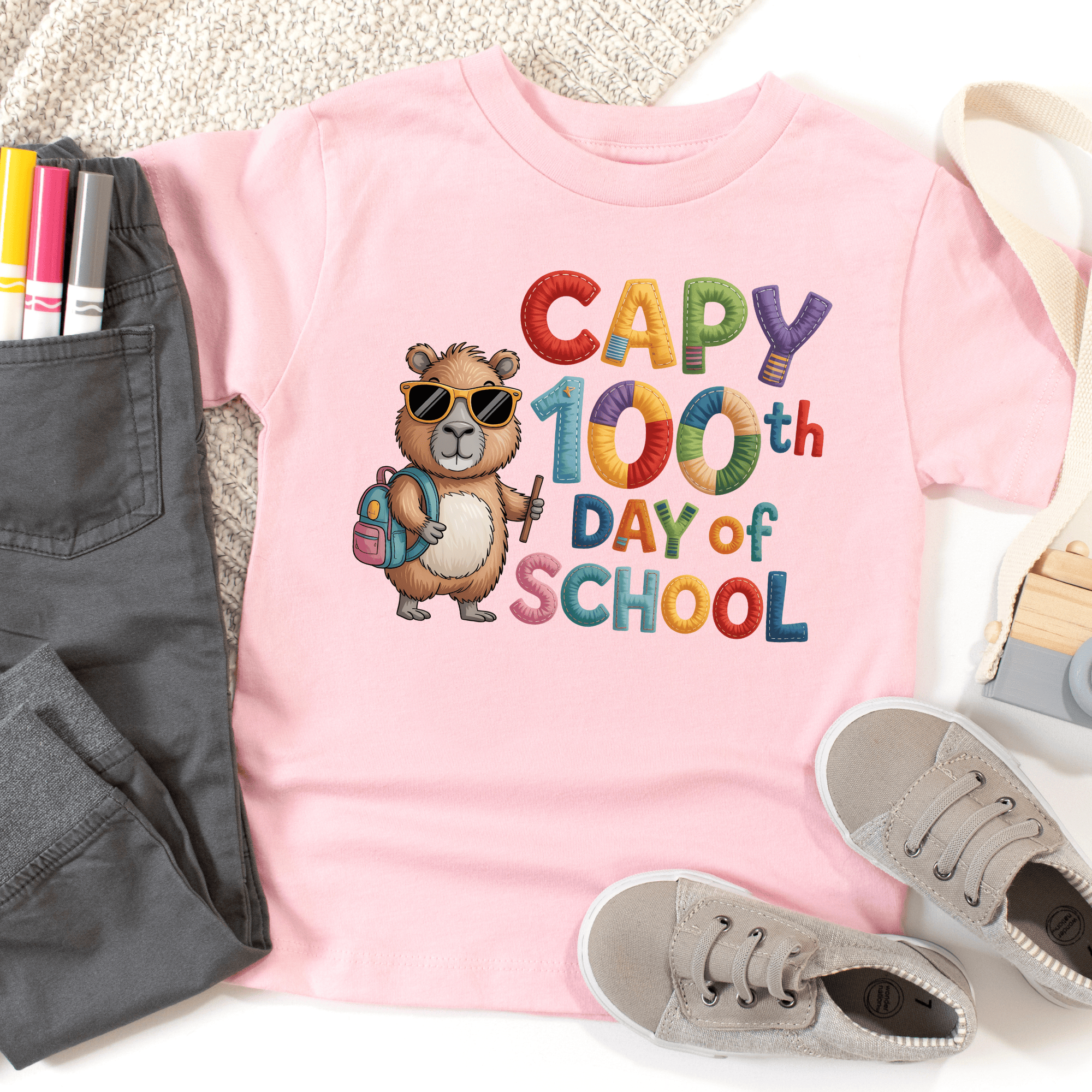 Pink t-shirt with a bear graphic and 'Capy 100th Day of School' text, surrounded by casual items.