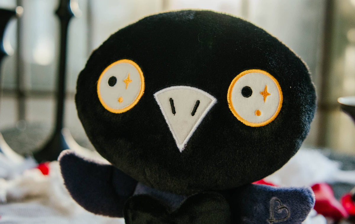 A black and blue raven plushie with a white beak and white and yellow eyes. Its wings are holding a black heart with the white text "Nevermore in love with you." The raven has yellow legs with a black "xoxo" on the right foot.