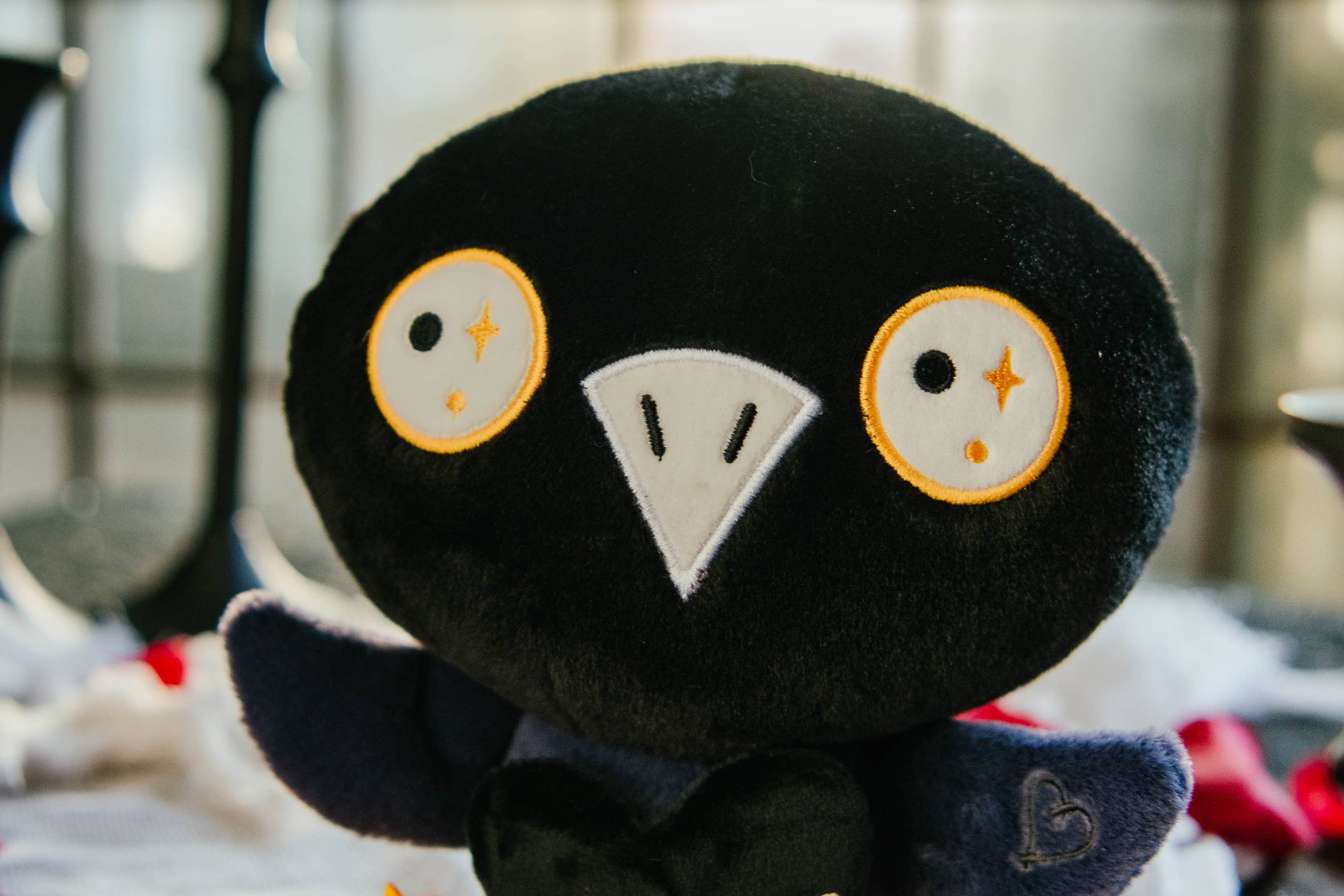 A black and blue raven plushie with a white beak and white and yellow eyes. Its wings are holding a black heart with the white text "Nevermore in love with you." The raven has yellow legs with a black "xoxo" on the right foot.