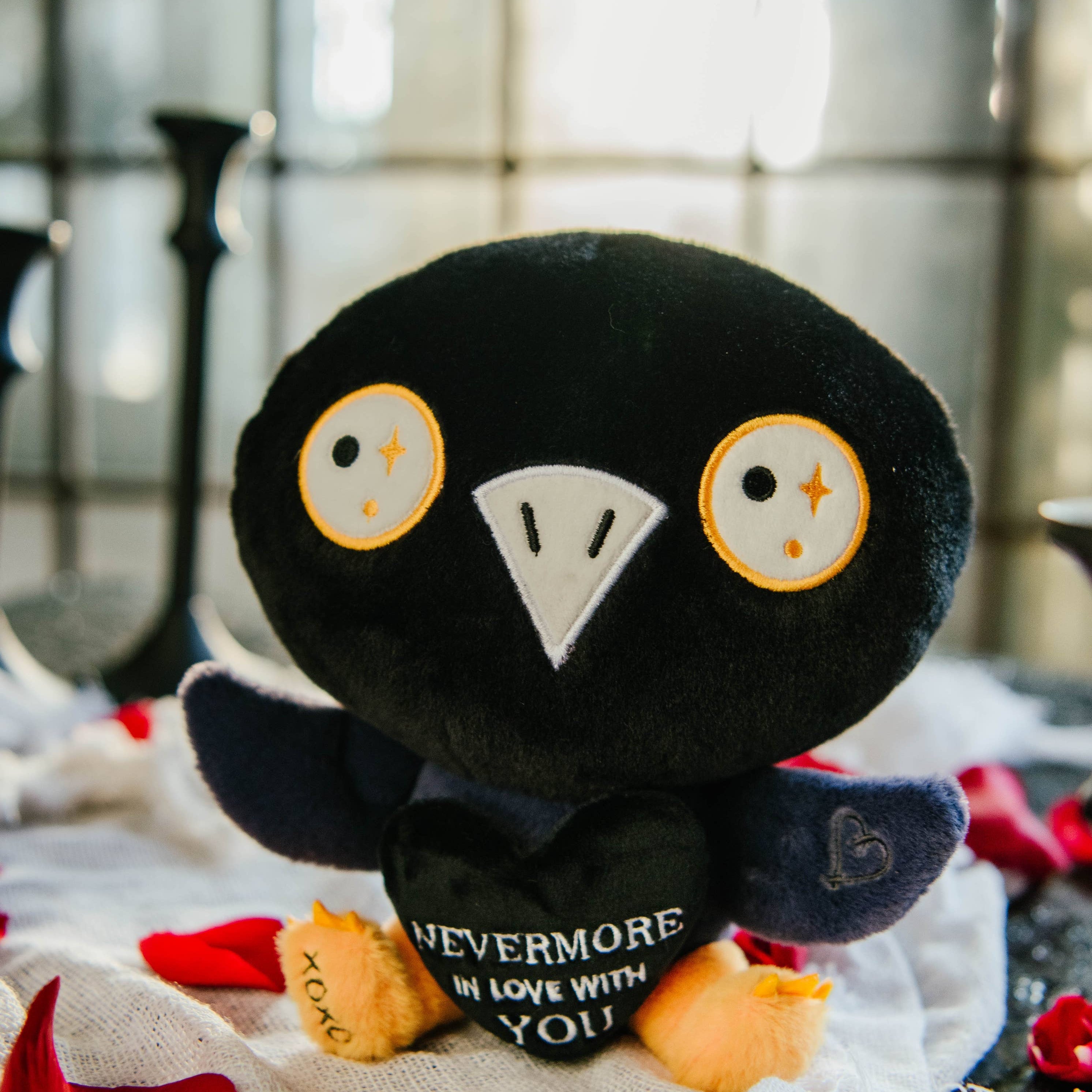 A black and blue raven plushie with a white beak and white and yellow eyes. Its wings are holding a black heart with the white text "Nevermore in love with you." The raven has yellow legs with a black "xoxo" on the right foot.