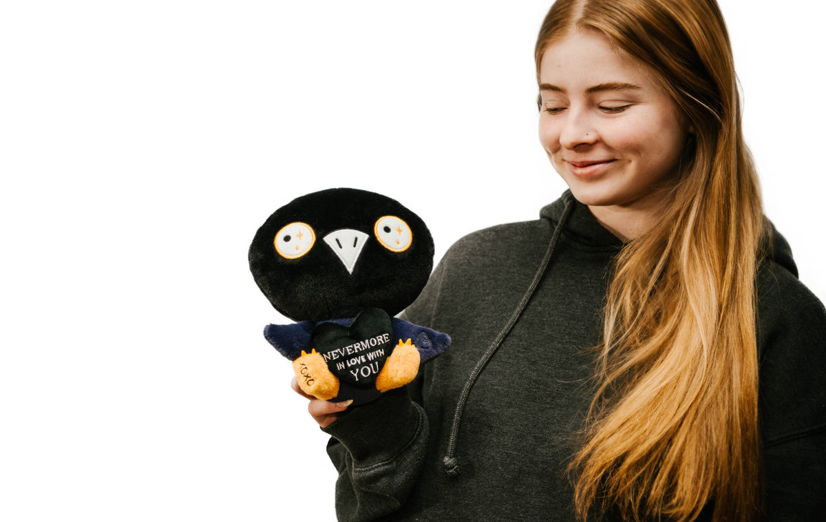 Woman holding a black and blue raven plushie with a white beak and white and yellow eyes. Its wings are holding a black heart with the white text "Nevermore in love with you." The raven has yellow legs with a black "xoxo" on the right foot.