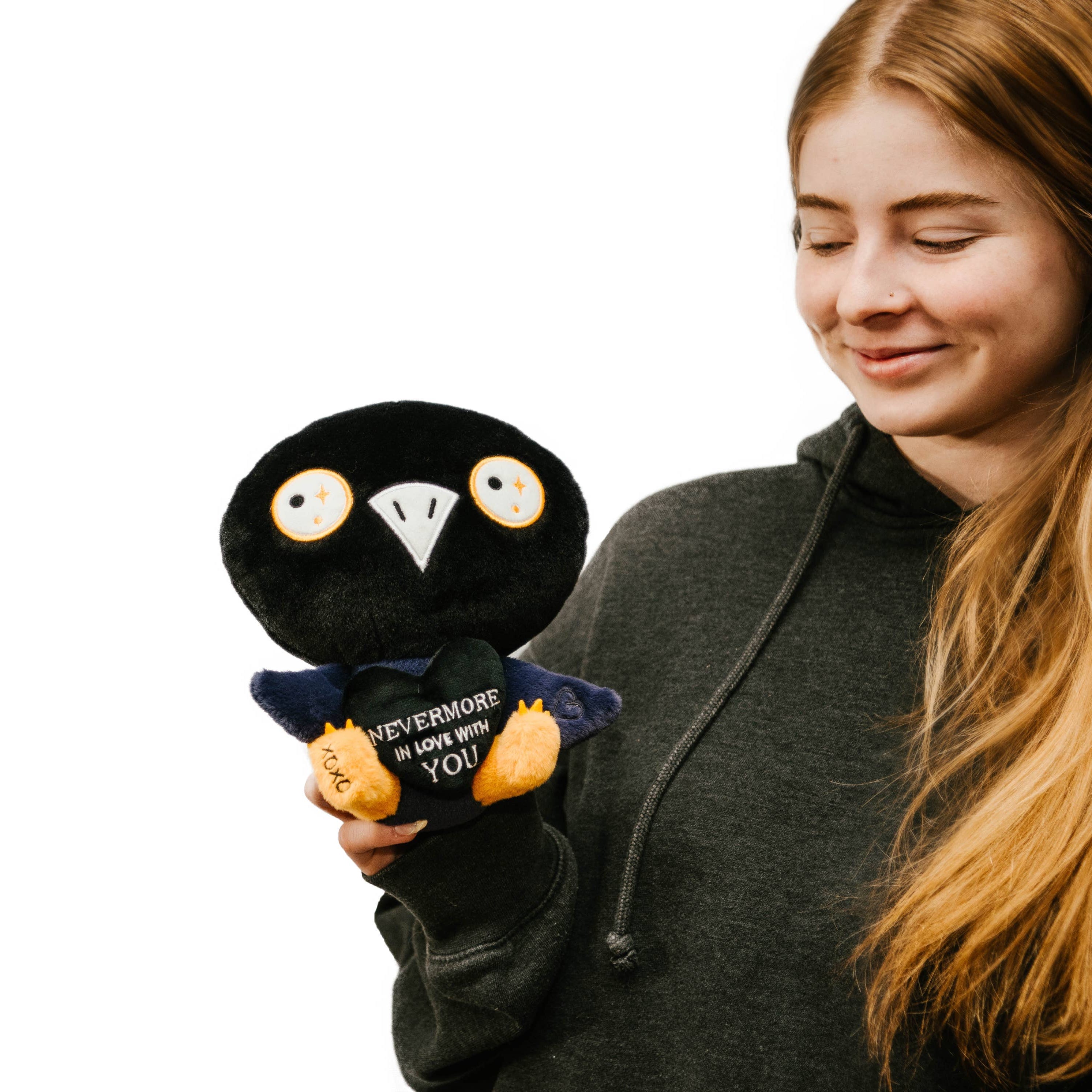 Woman holding a black and blue raven plushie with a white beak and white and yellow eyes. Its wings are holding a black heart with the white text "Nevermore in love with you." The raven has yellow legs with a black "xoxo" on the right foot.