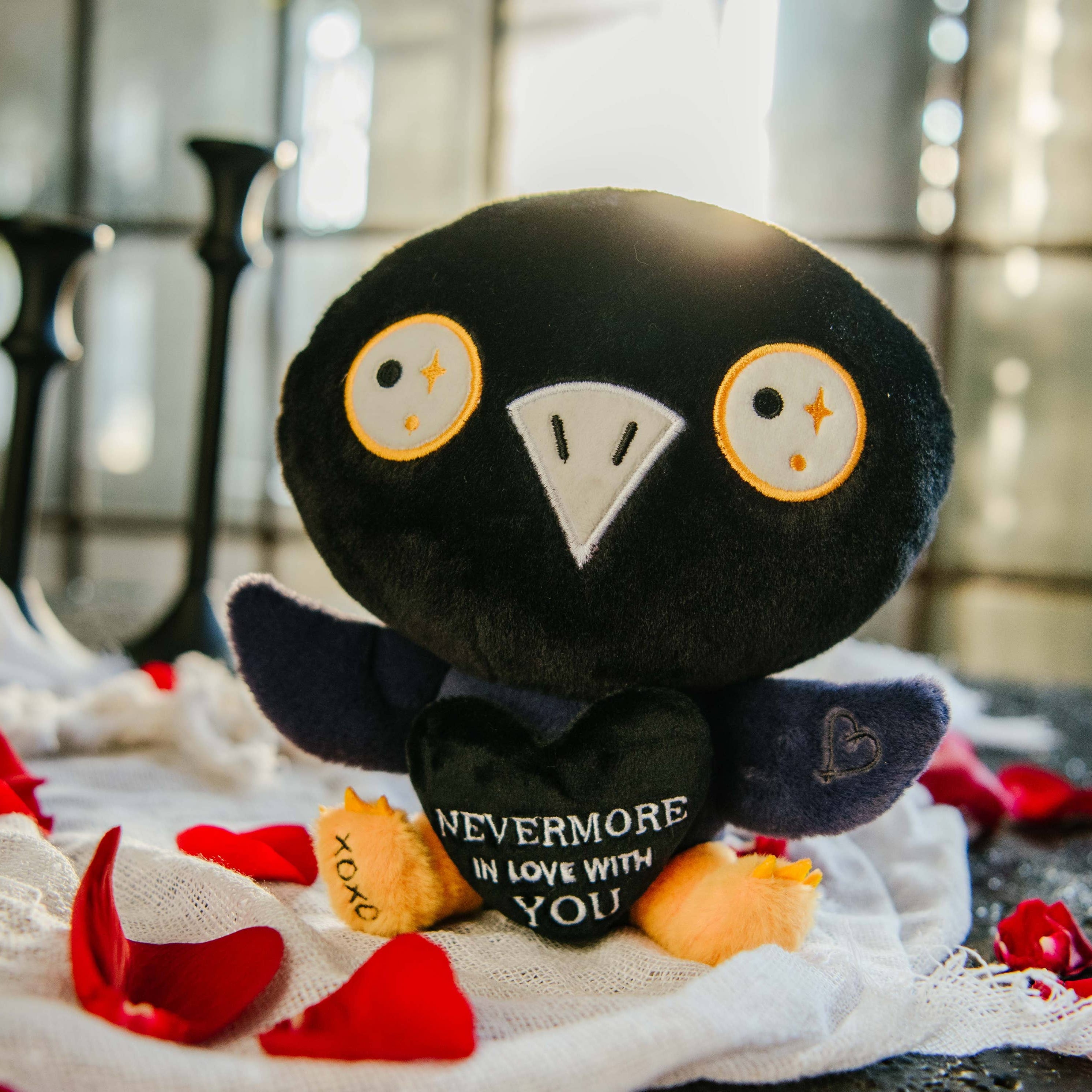 A black and blue raven plushie with a white beak and white and yellow eyes. Its wings are holding a black heart with the white text "Nevermore in love with you." The raven has yellow legs with a black "xoxo" on the right foot.