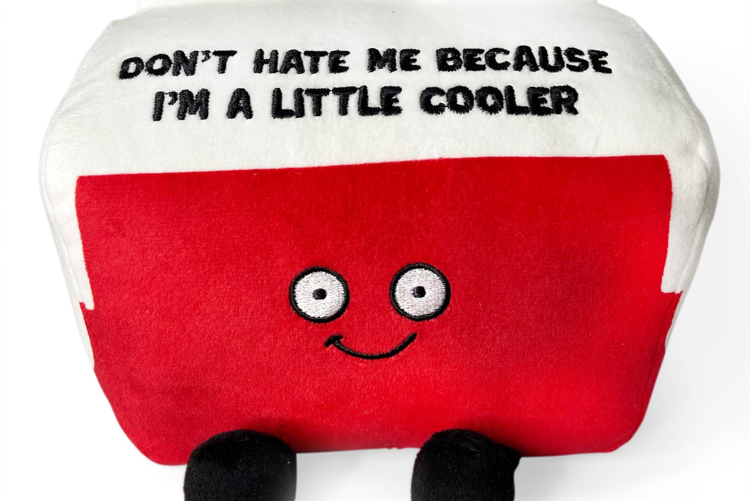 A cooler plushie with a white top and red bottom. The white top features black embroidered text that says, "Don't hate me because I'm a little cooler." The red section has a smiling face, with black legs sticking out from the bottom.