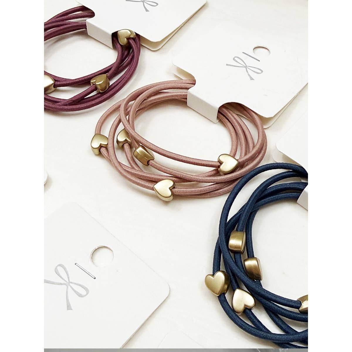 Three sets of elastic hair ties in pink, maroon, and blue, each adorned with gold heart charms. They rest on white cards with a small loop.