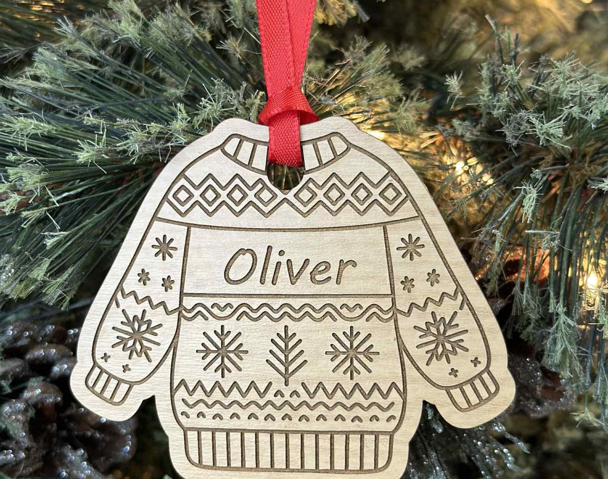 Wooden ornament shaped like a sweater with 'Oliver' on it, hanging on a Christmas tree.