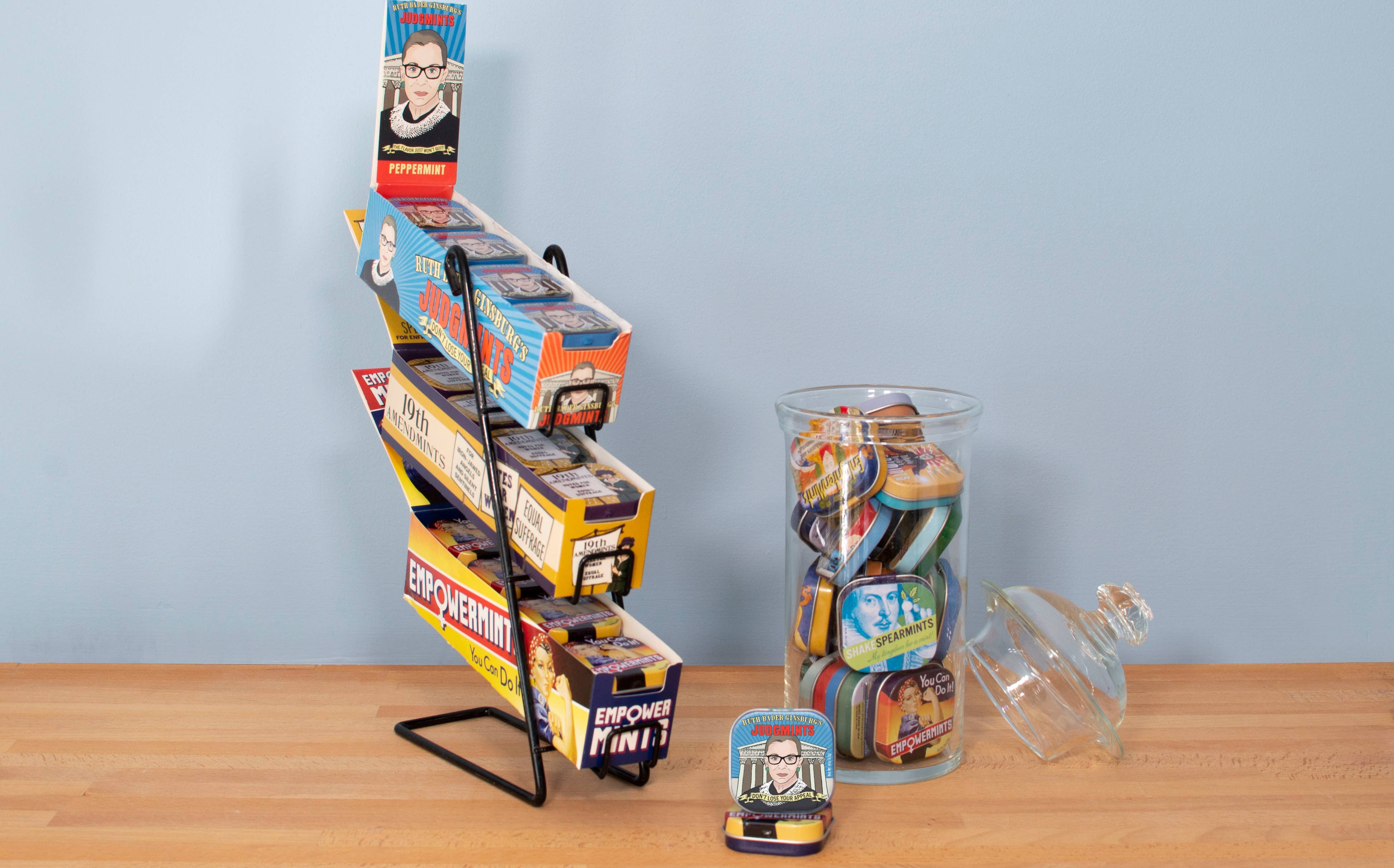 Display stand of colorful mint tins with empowering themes and graphics. A jar beside it is filled with similar tins. Light and uplifting tone.