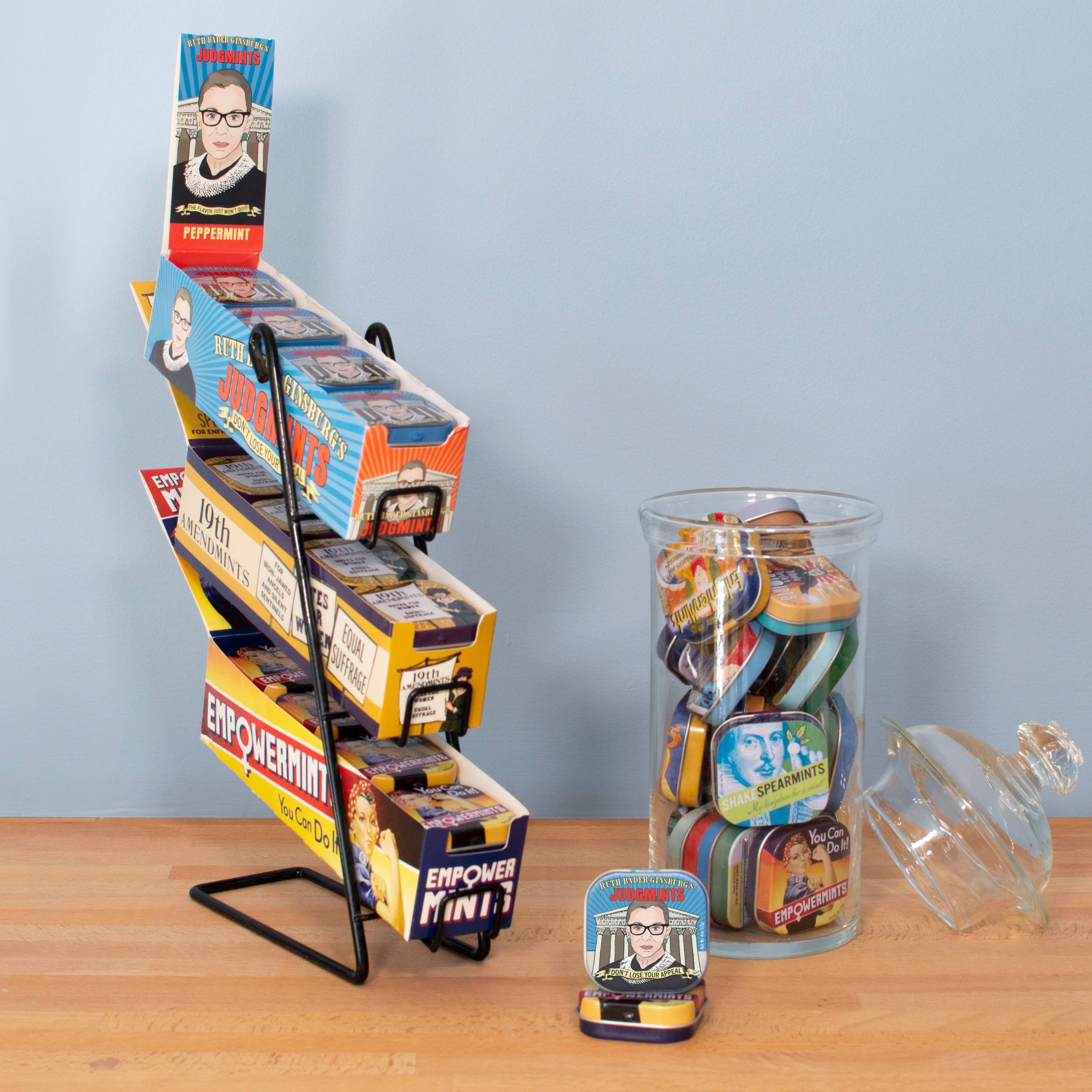 Display stand of colorful mint tins with empowering themes and graphics. A jar beside it is filled with similar tins. Light and uplifting tone.