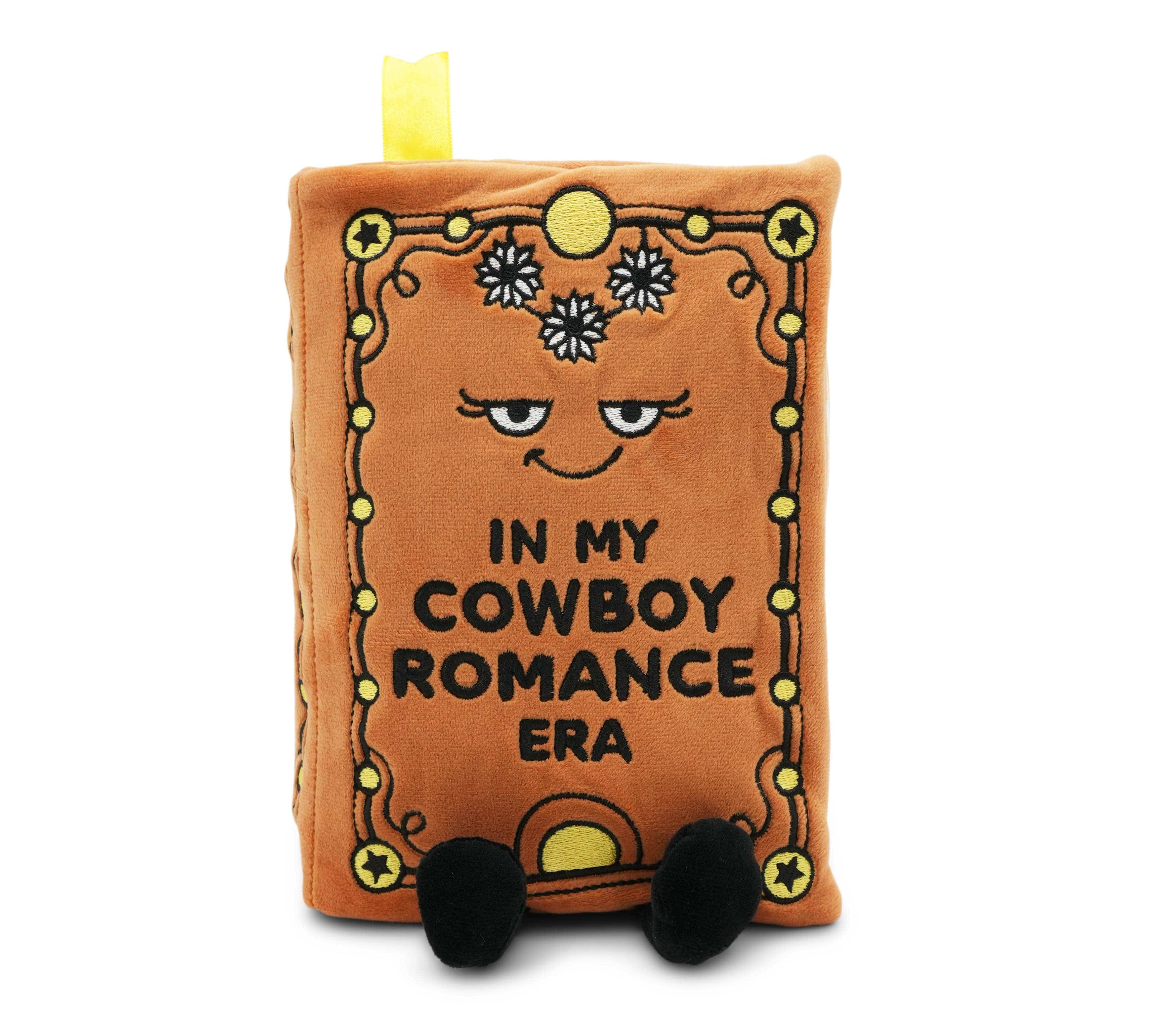 Plush toy resembling a book with a face and text 'In my cowboy romance era' on a white background