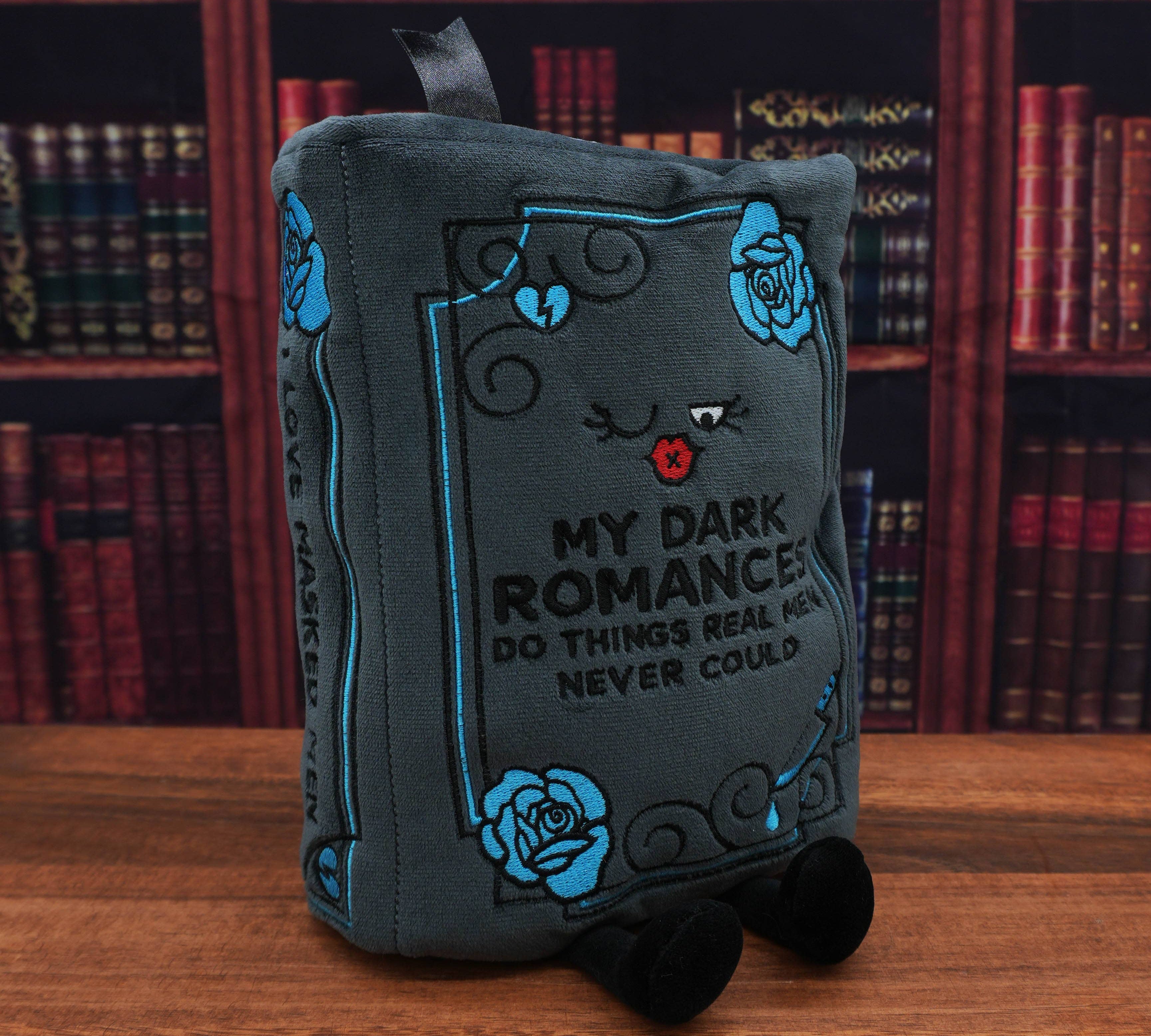 Plush book-shaped pillow with decorative text and roses on a wooden surface with bookshelf background