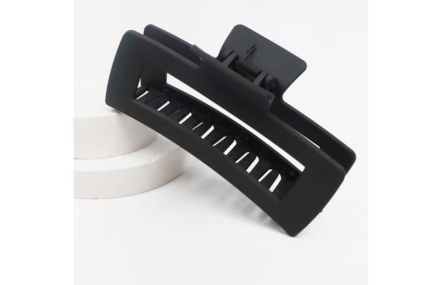 A large, matte black jumbo rectangular hair claw clip with a hollow cut-out design, propped on a white circular display platform.