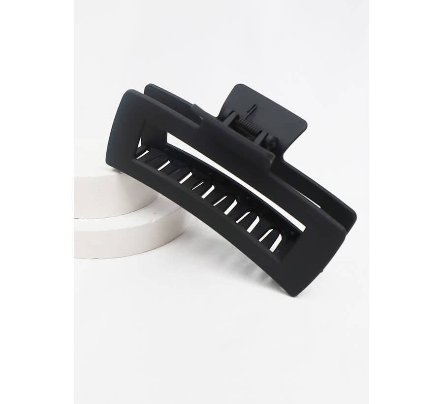 A large, matte black jumbo rectangular hair claw clip with a hollow cut-out design, propped on a white circular display platform.