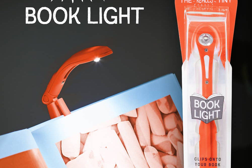 A small, orange book light is clipped to an open book, illuminating the pages. Packaging on the right showcases "The Really Tiny Book Light" branding.