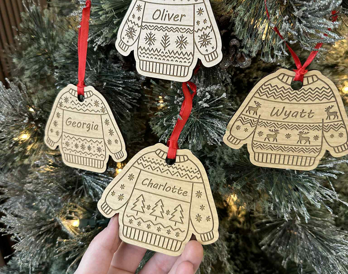 Decorative Christmas ornaments shaped like sweaters and mittens with names on a Christmas tree.
