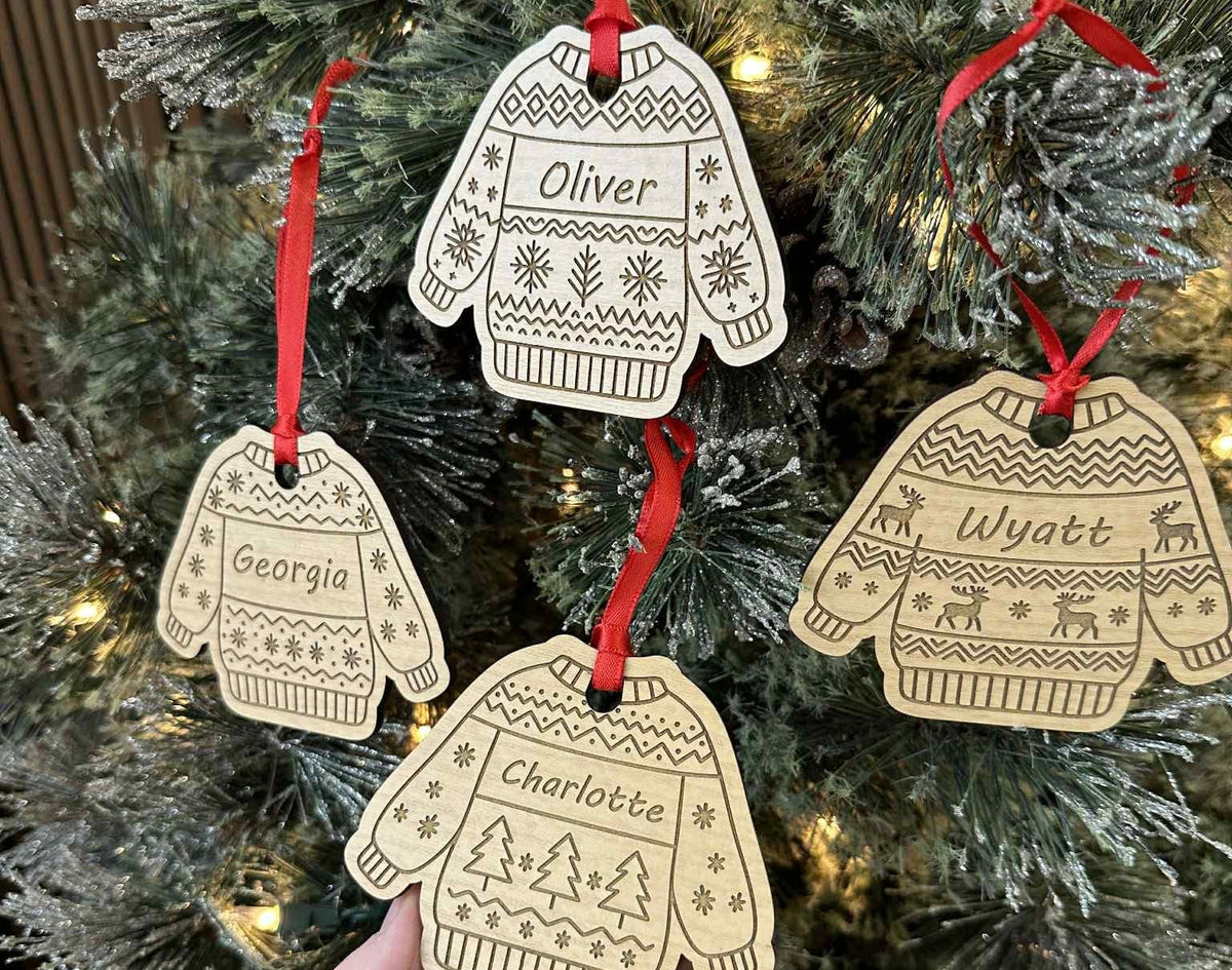Decorative Christmas ornaments shaped like sweaters and mittens with names on a tree.