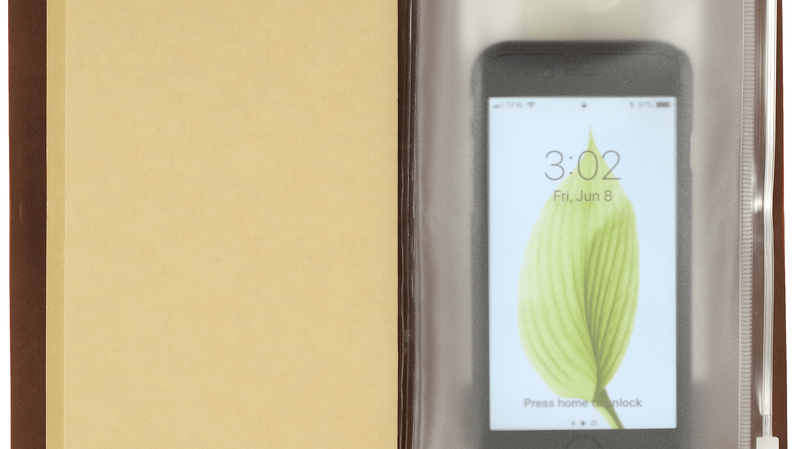 Open notebook with a beige page on the left and a smartphone inside a clear plastic sleeve on the right. The phone displays a leaf on its lock screen.