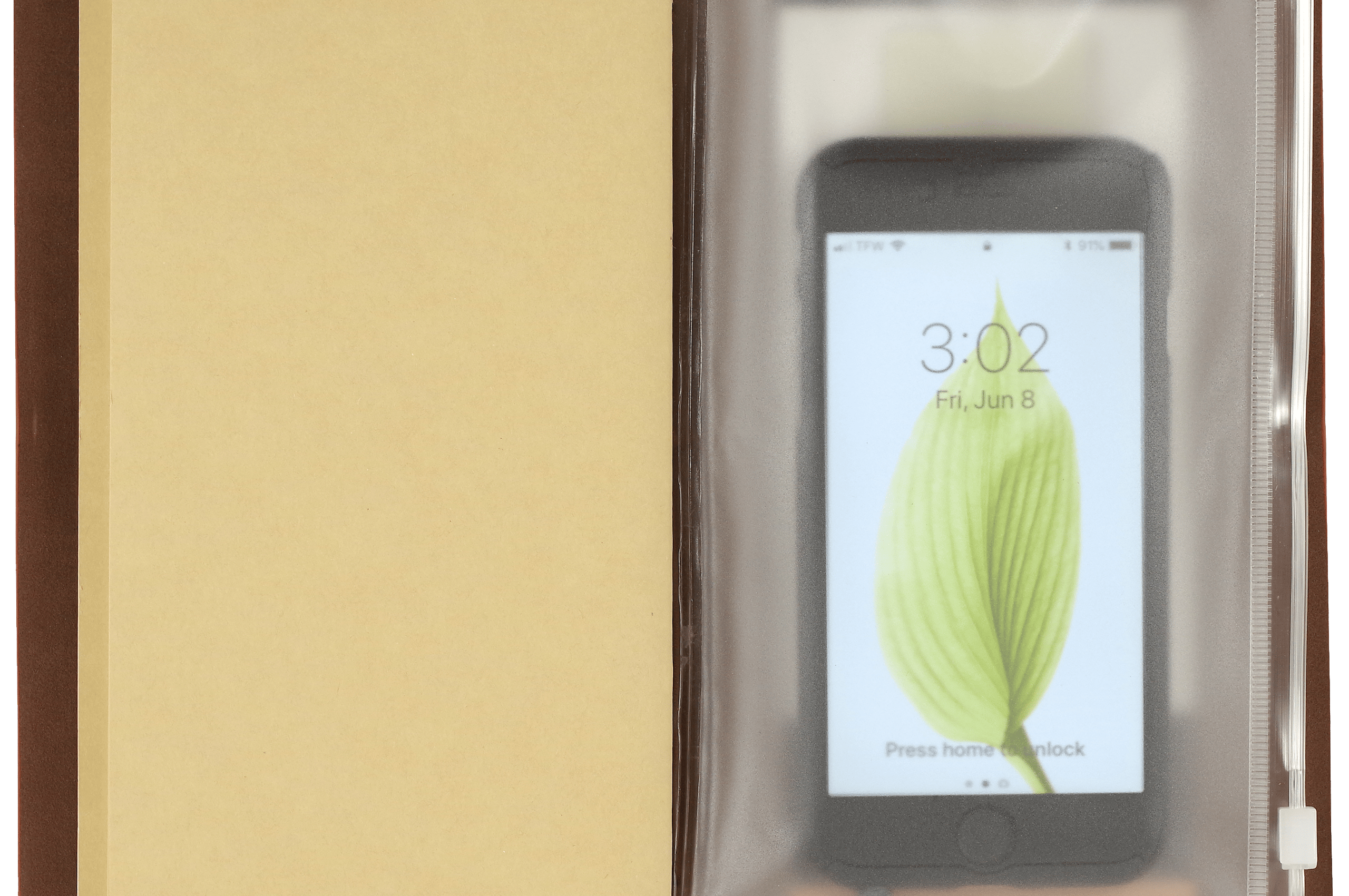 Open notebook with a beige page on the left and a smartphone inside a clear plastic sleeve on the right. The phone displays a leaf on its lock screen.