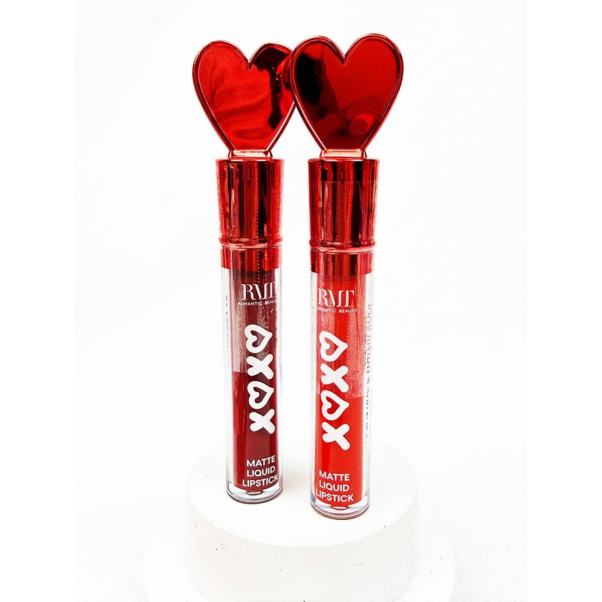 Two tubes of red matte liquid lipstick with shiny heart-shaped caps stand on a white pedestal. The packaging is sleek and vibrant, evoking a romantic tone.