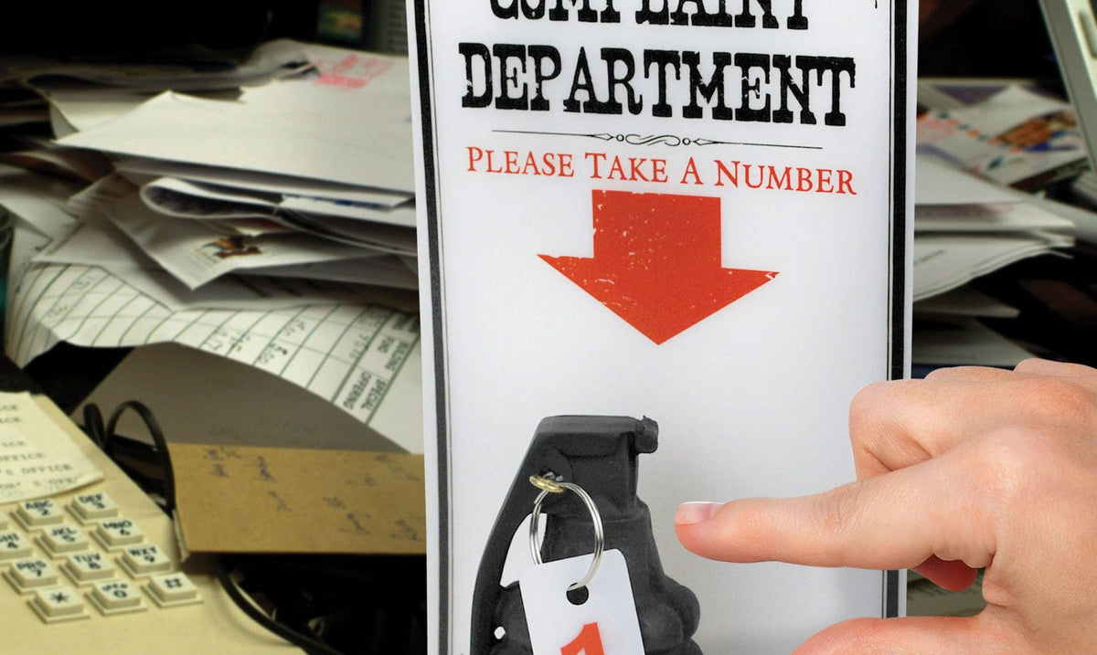 A hand reaches for a number tag attached to a grenade beneath a sign reading "Complaint Department, Please Take a Number," set on a cluttered desk.