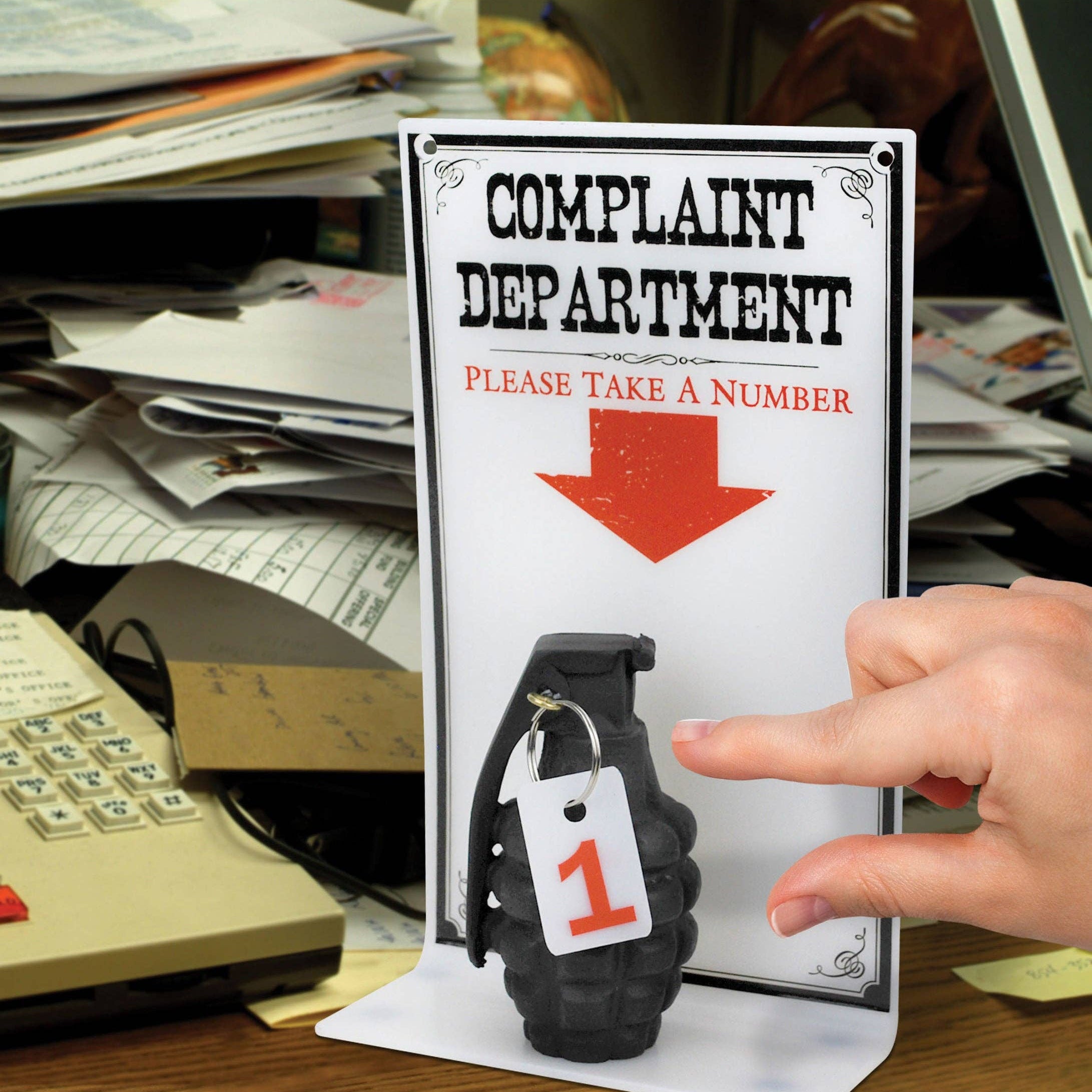 A hand reaches for a number tag attached to a grenade beneath a sign reading "Complaint Department, Please Take a Number," set on a cluttered desk.