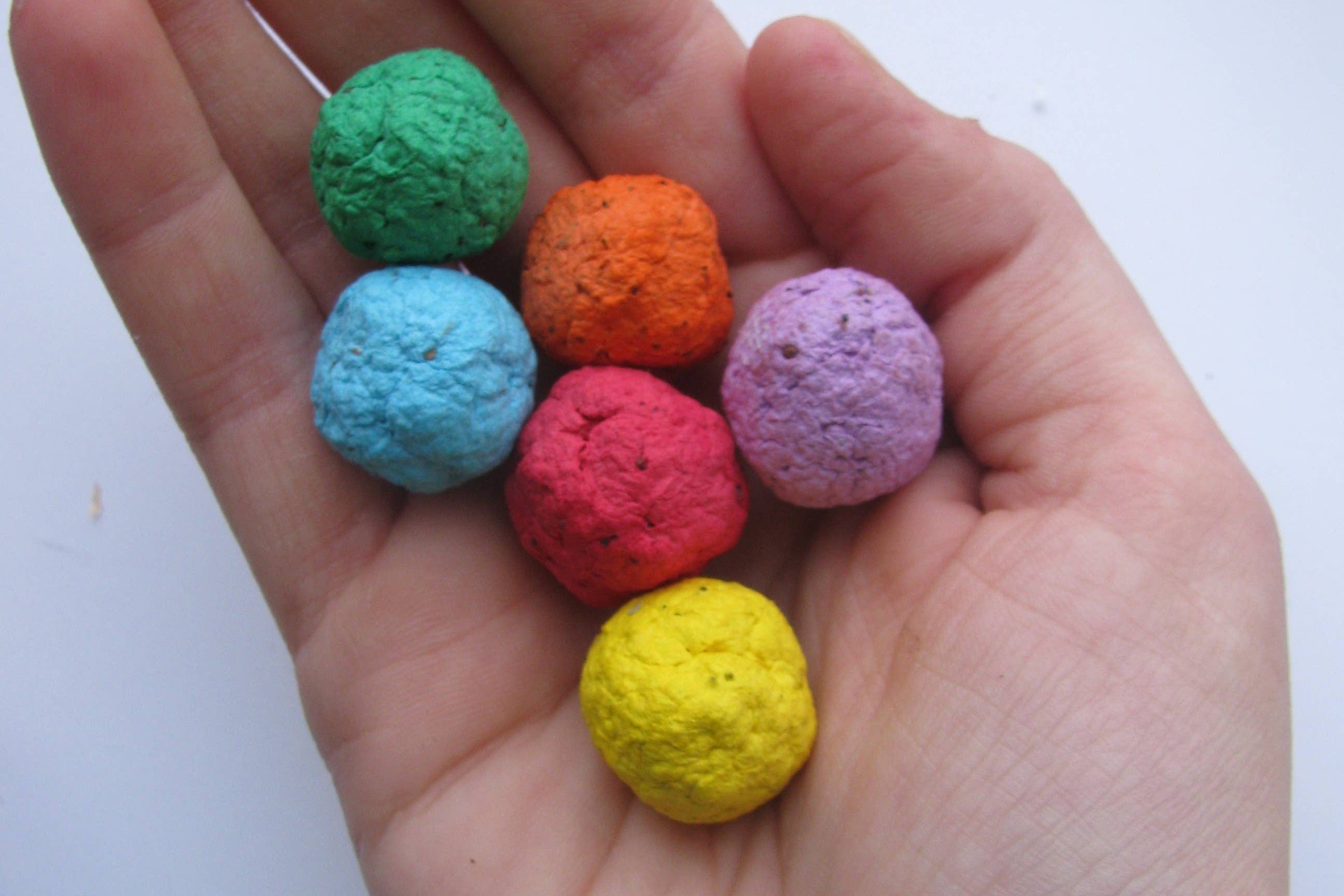 A hand holds six small, round seed bombs, each a different color: green, orange, blue, purple, red, and yellow, on a white background. Colorful and inviting.
