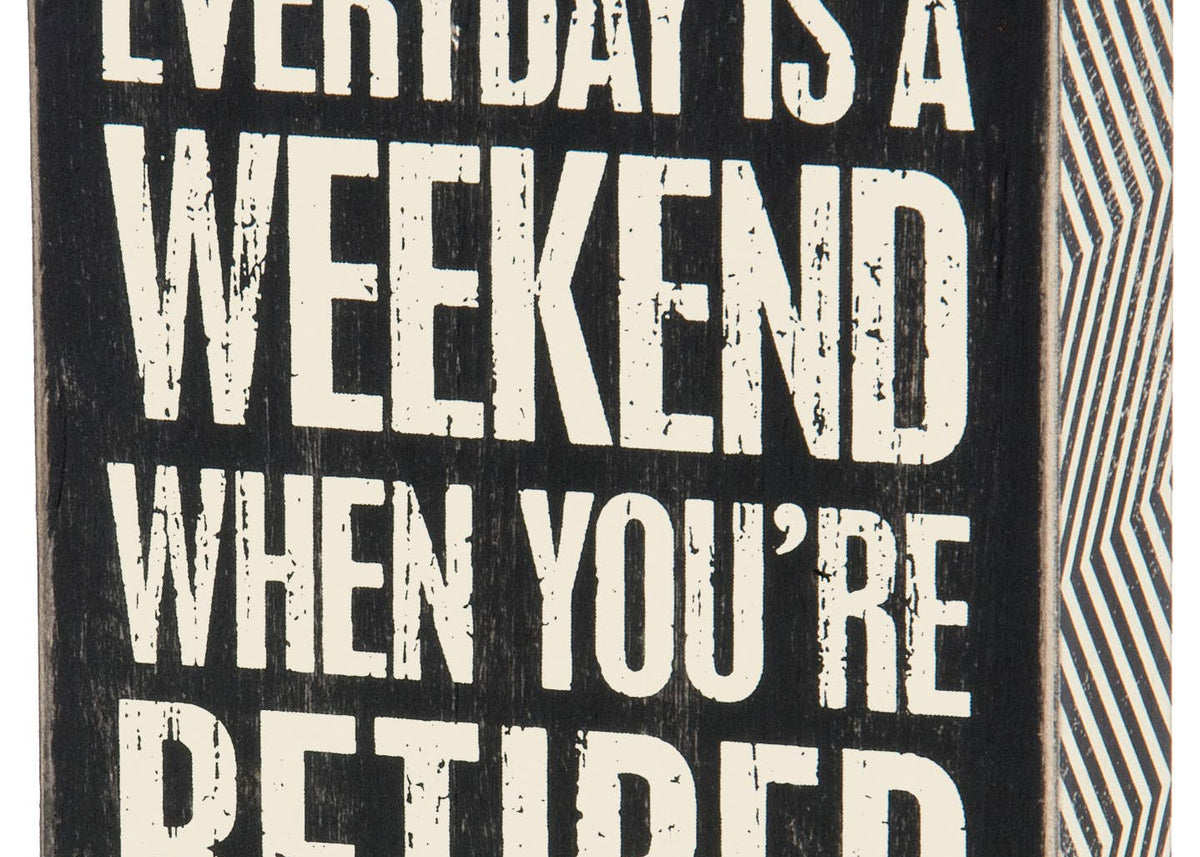 A square black wooden box sign with white capitalized text that reads 'EVERYDAY IS A WEEKEND WHEN YOU’RE RETIRED.' The sign features a distressed vintage finish and a decorative black and white zigzag pattern on the side panels.