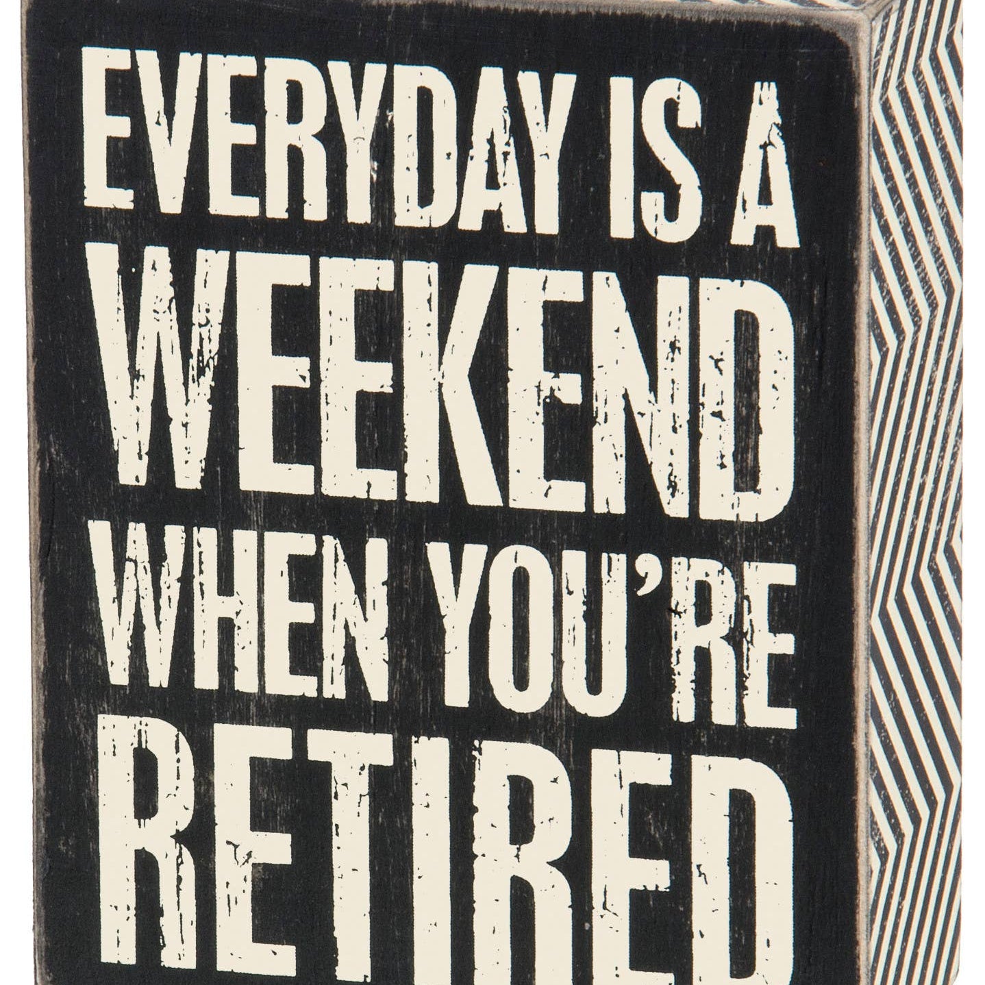 A square black wooden box sign with white capitalized text that reads 'EVERYDAY IS A WEEKEND WHEN YOU’RE RETIRED.' The sign features a distressed vintage finish and a decorative black and white zigzag pattern on the side panels.