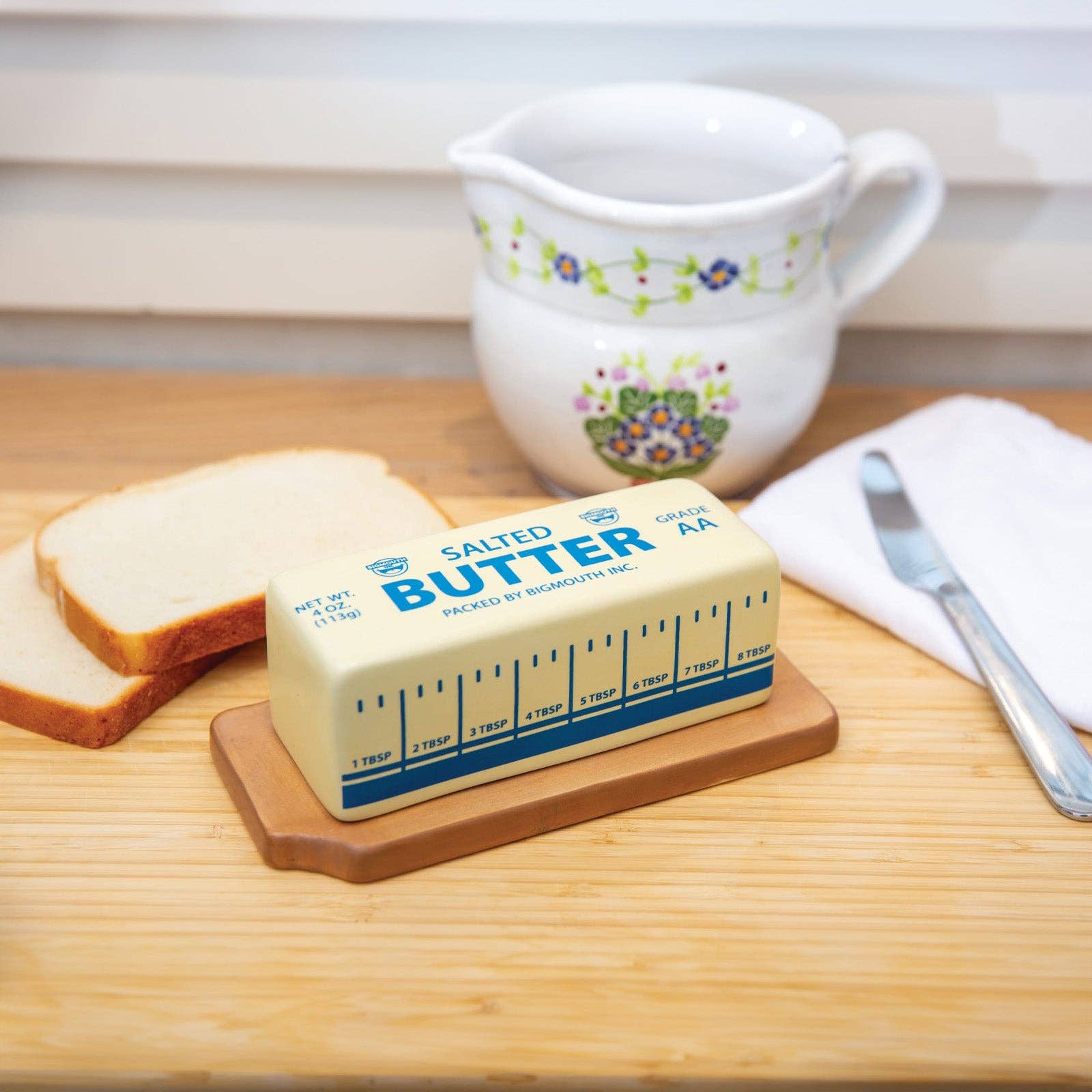 Stick of salted butter on a wooden tray beside sliced bread, a floral ceramic jug, a butter knife, and a white napkin. Cozy kitchen setting.