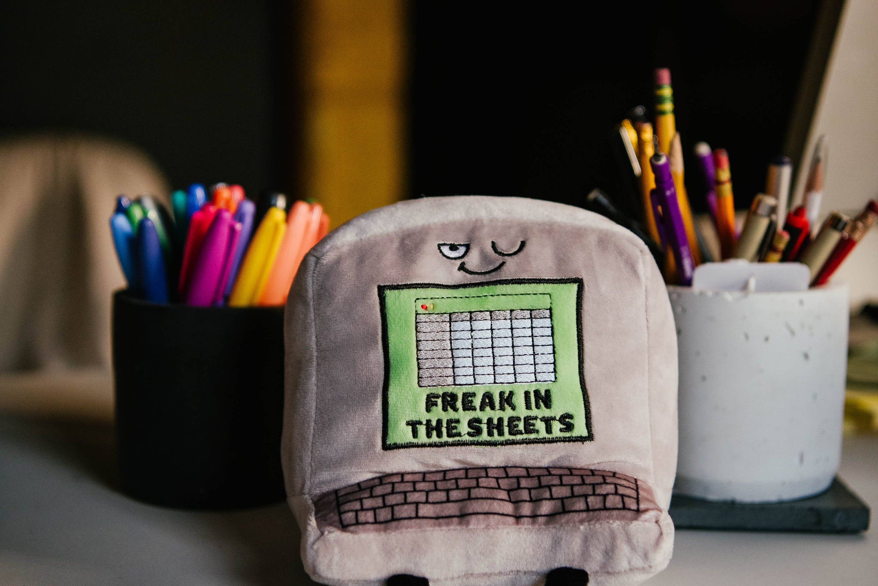 A gray computer plush with a winky face, featuring a green Excel-style sheet embroidered with the words "Freak in the Sheets" in black. It also has an outlined keyboard design and black legs sticking out from the bottom. Plush is sitting on a desk.