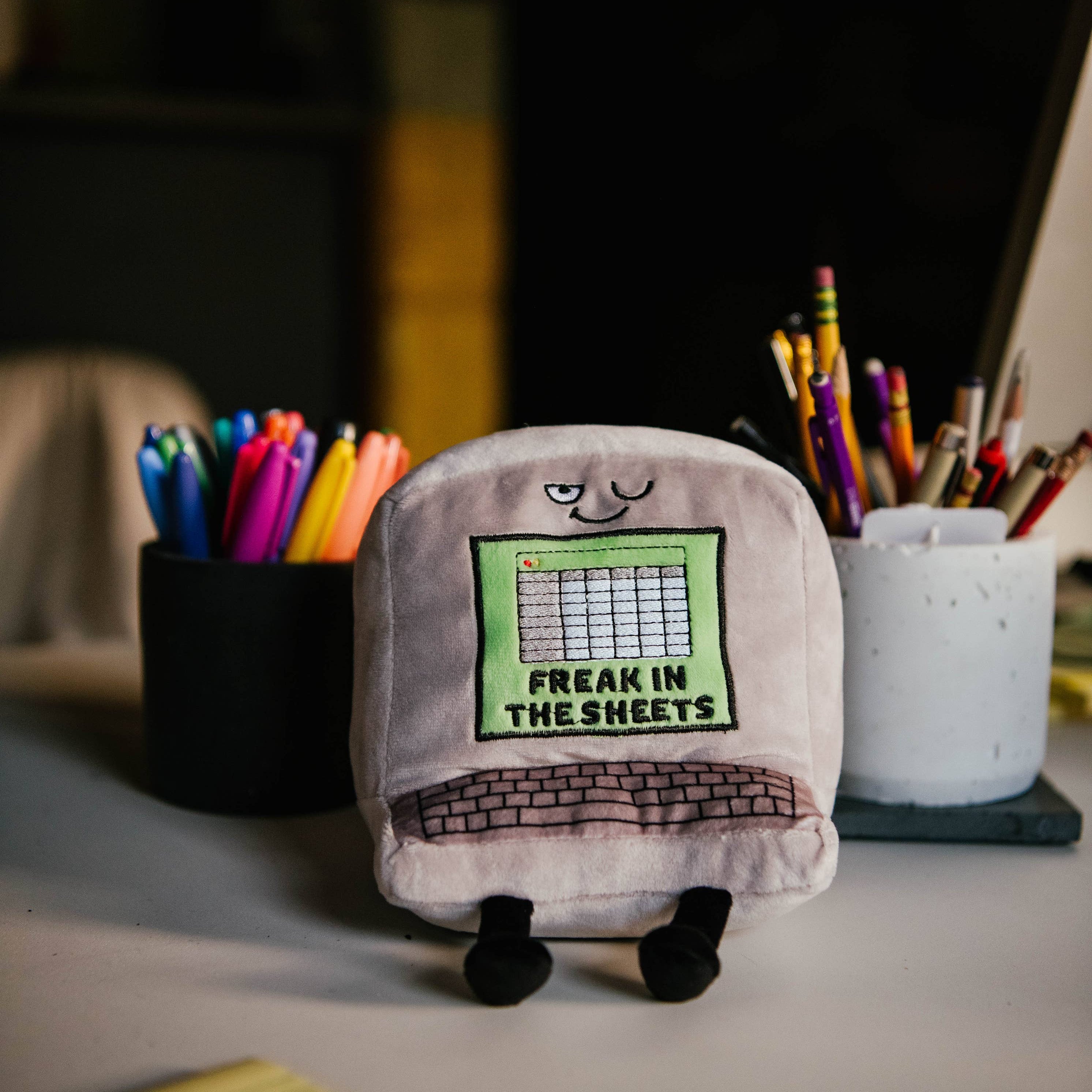 A gray computer plush with a winky face, featuring a green Excel-style sheet embroidered with the words "Freak in the Sheets" in black. It also has an outlined keyboard design and black legs sticking out from the bottom. Plush is sitting on a desk.