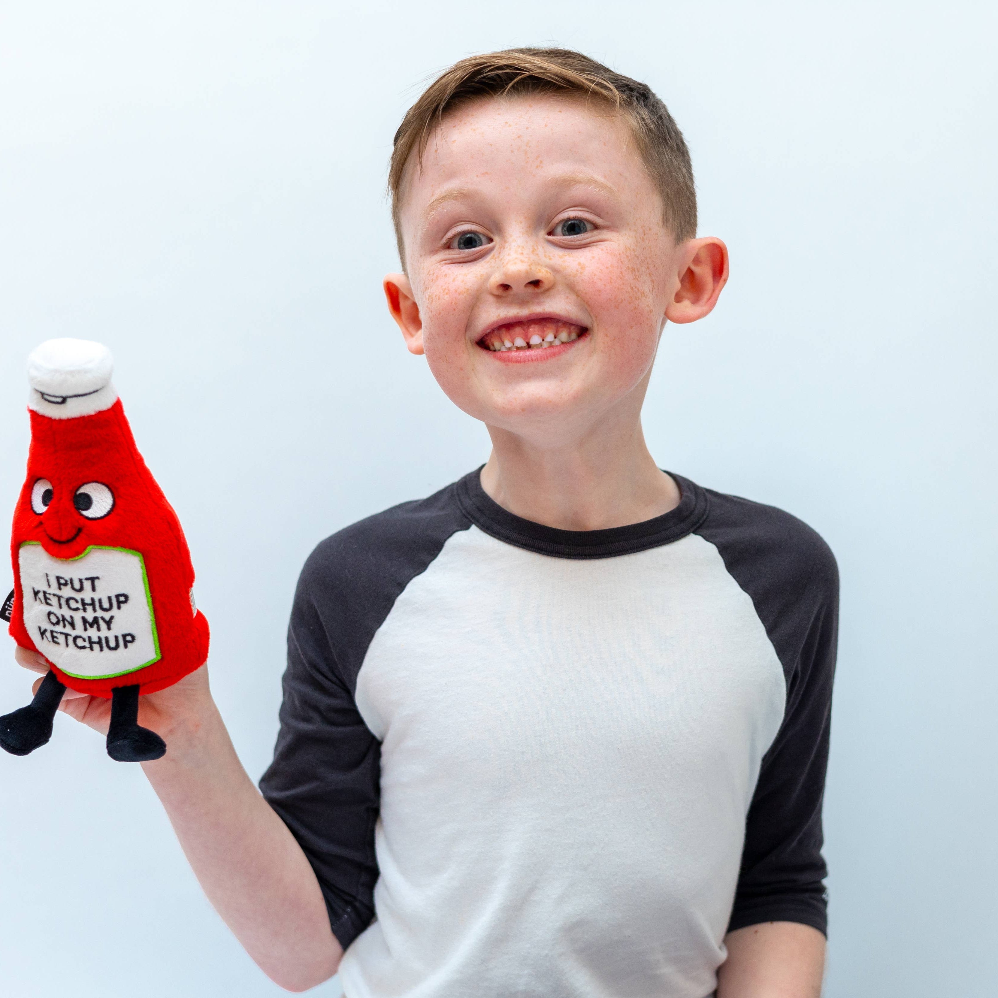 Child holding a ketchup bottle plushie with a smiley face and a white belly featuring black text that says "I put ketchup on my ketchup." It has a white lid and black legs sticking out.