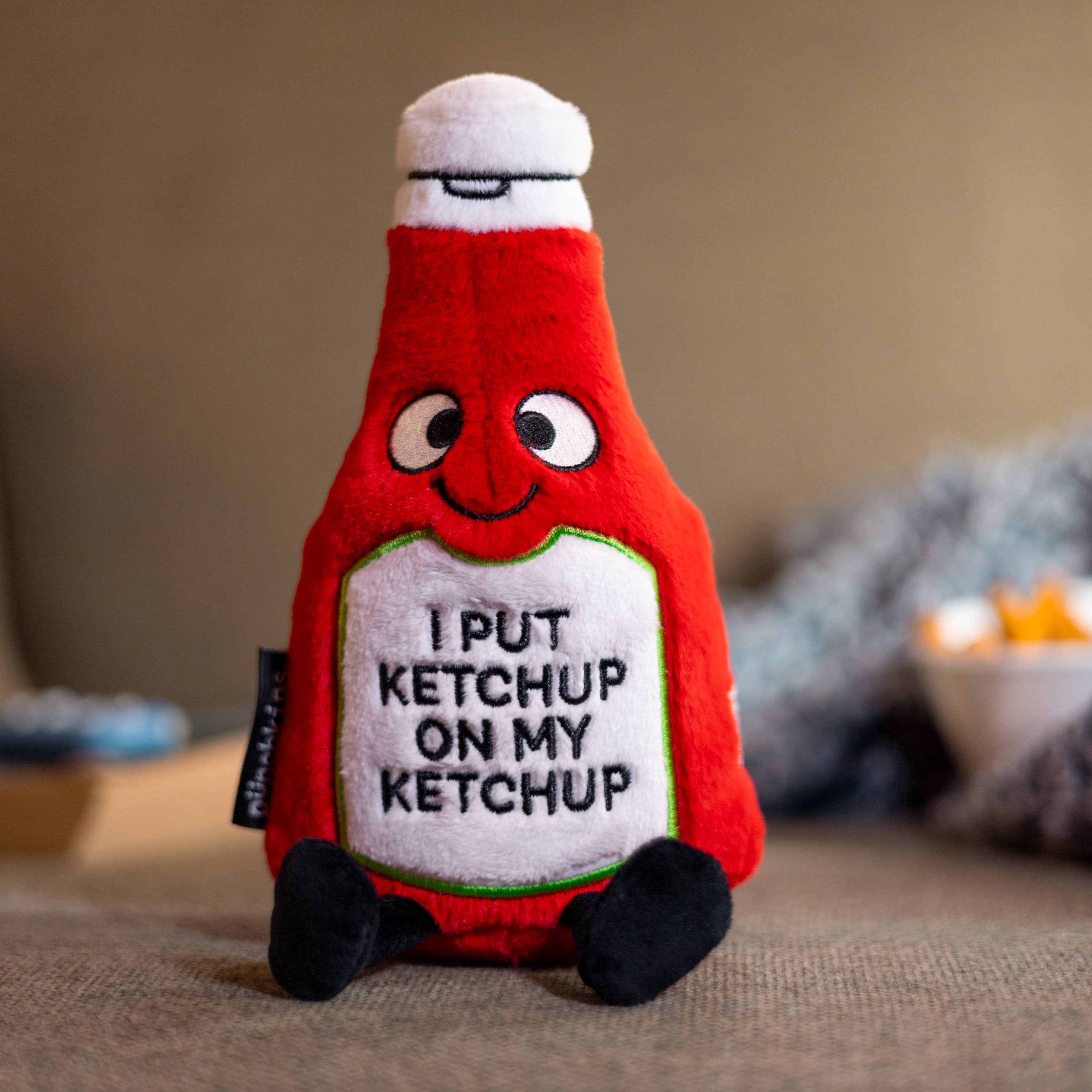 A ketchup bottle plushie with a smiley face and a white belly featuring black text that says "I put ketchup on my ketchup." It has a white lid and black legs sticking out.