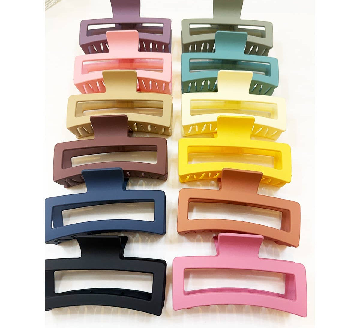 An assortment of twelve large, matte rectangular hair claw clips with hollow cut-out designs, arranged in two vertical rows showing a variety of pastel and neutral colors including pink, green, yellow, and navy blue.
