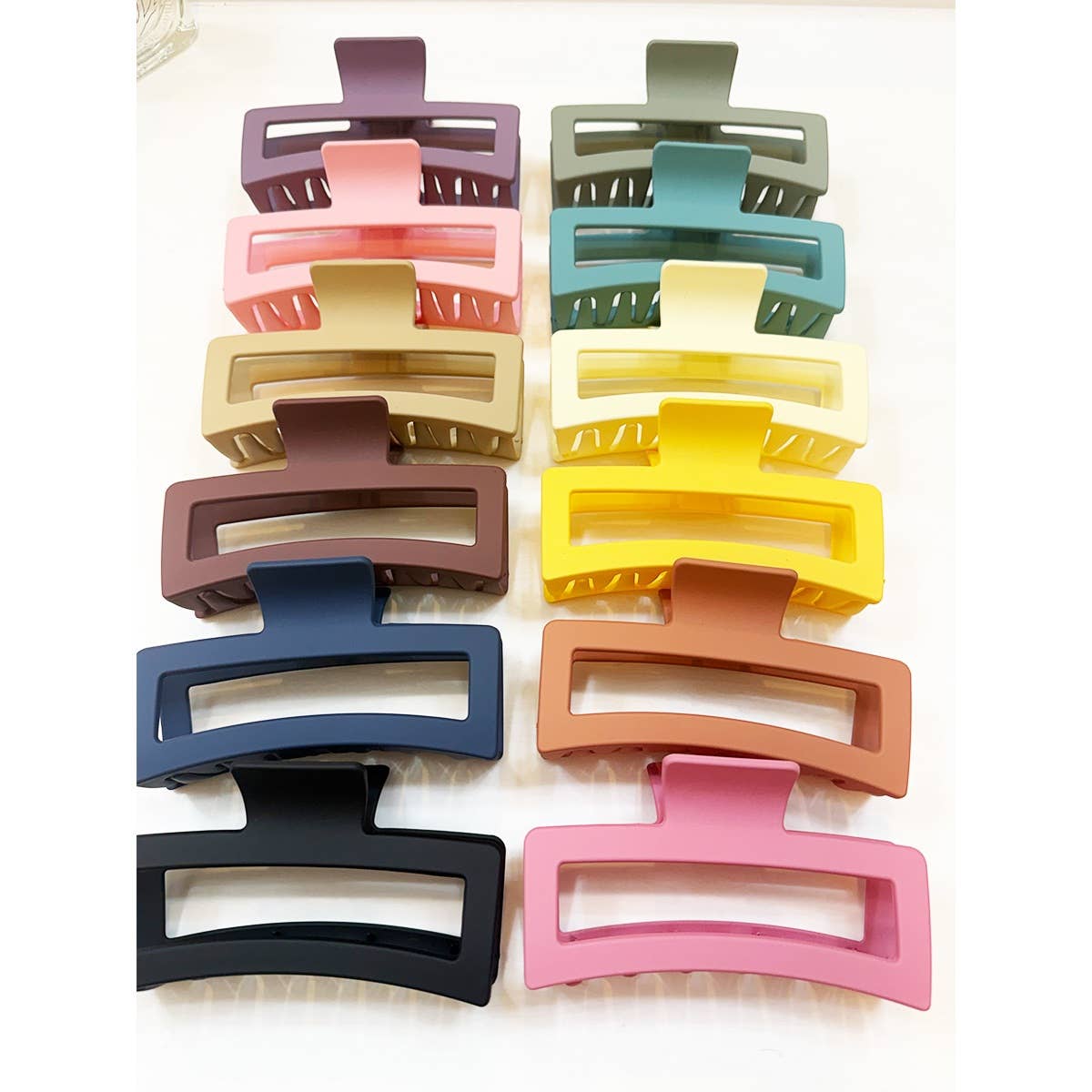 An assortment of twelve large, matte rectangular hair claw clips with hollow cut-out designs, arranged in two vertical rows showing a variety of pastel and neutral colors including pink, green, yellow, and navy blue.