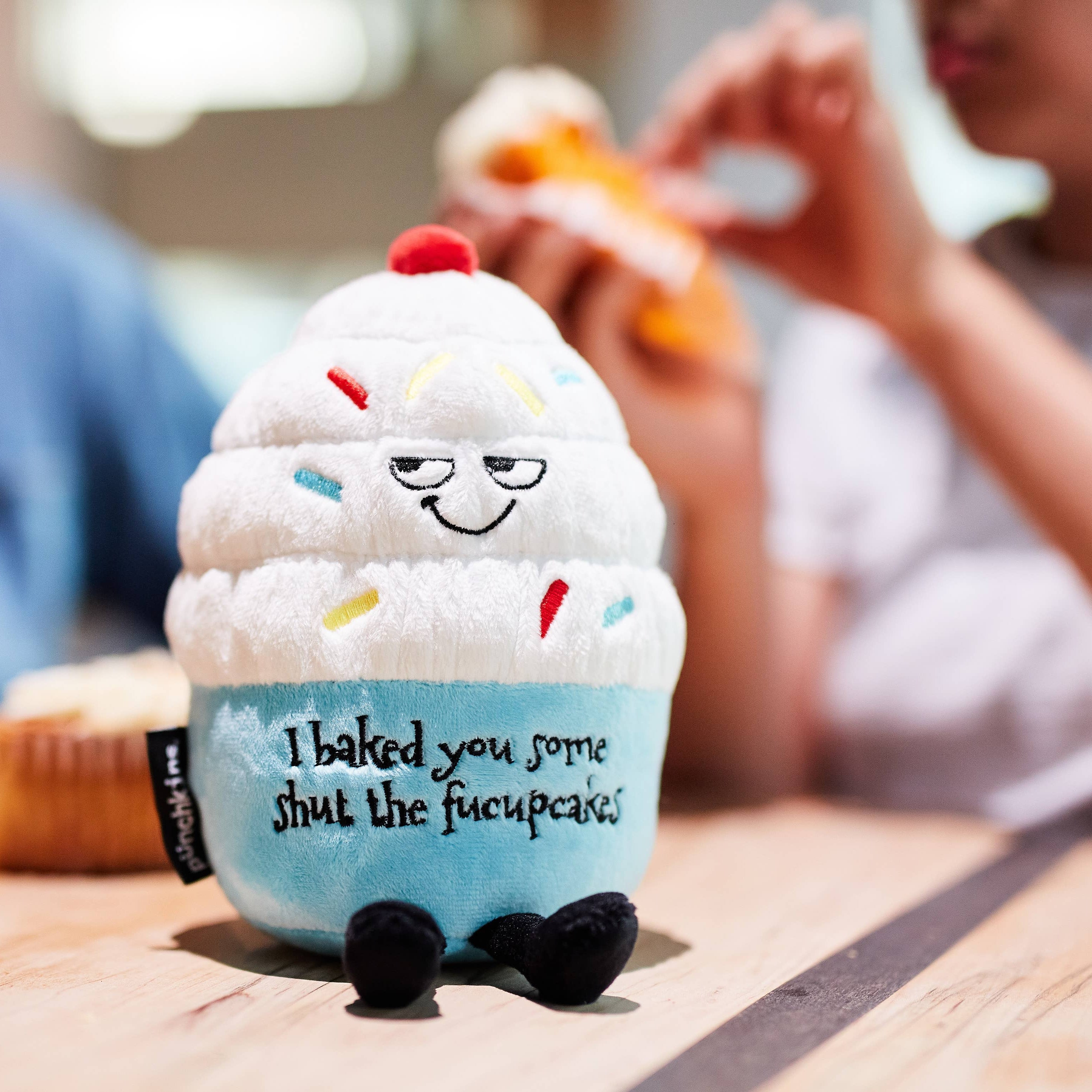A cupcake plush with white frosting, rainbow sprinkles, and a blue base. The top features a sly, smiling face, while the bottom has black embroidered text that reads, "I baked you some shut the fucupcakes." Black legs sticking out the bottom and a red cherry on top.