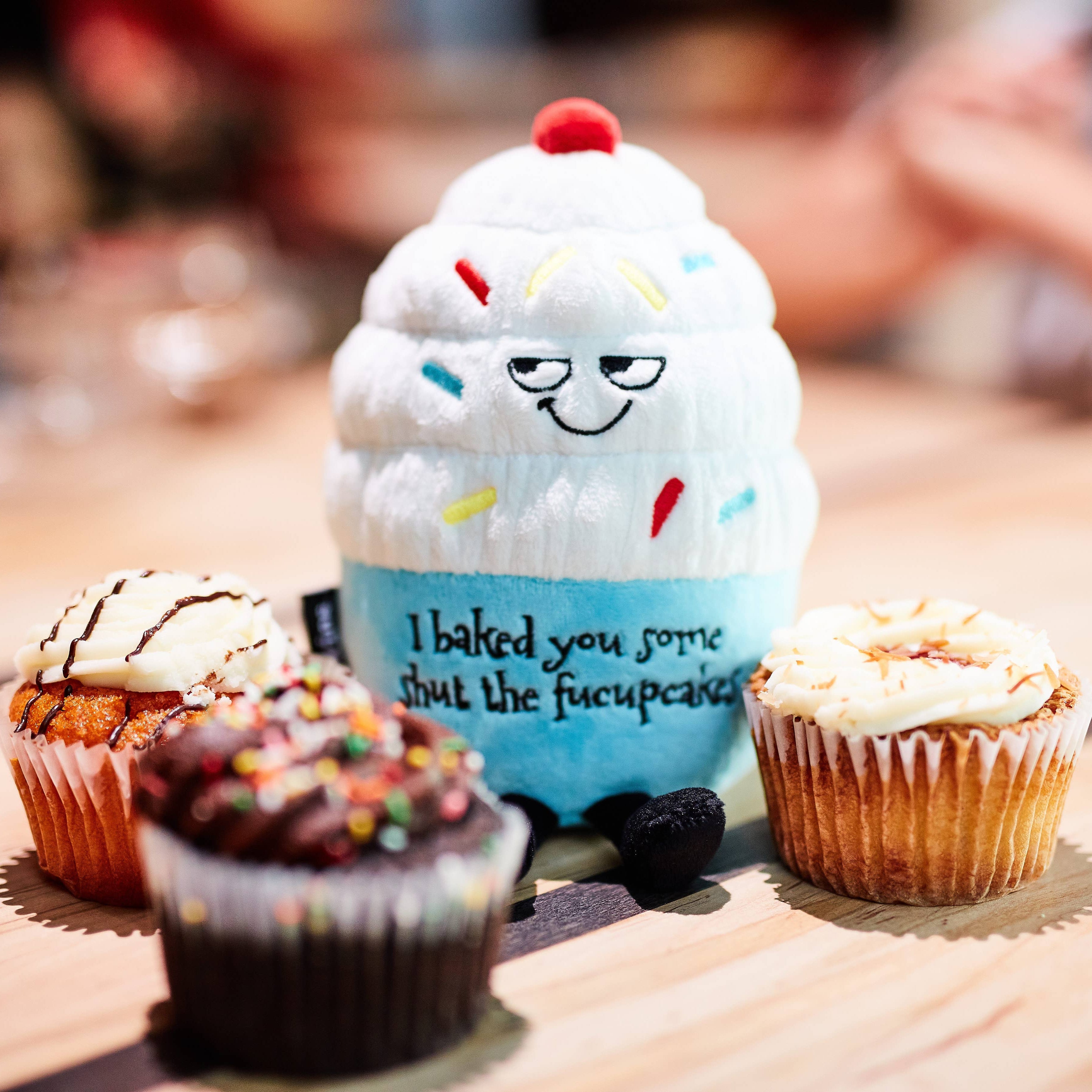 A cupcake plush with white frosting, rainbow sprinkles, and a blue base. The top features a sly, smiling face, while the bottom has black embroidered text that reads, "I baked you some shut the fucupcakes." Black legs sticking out the bottom and a red cherry on top.