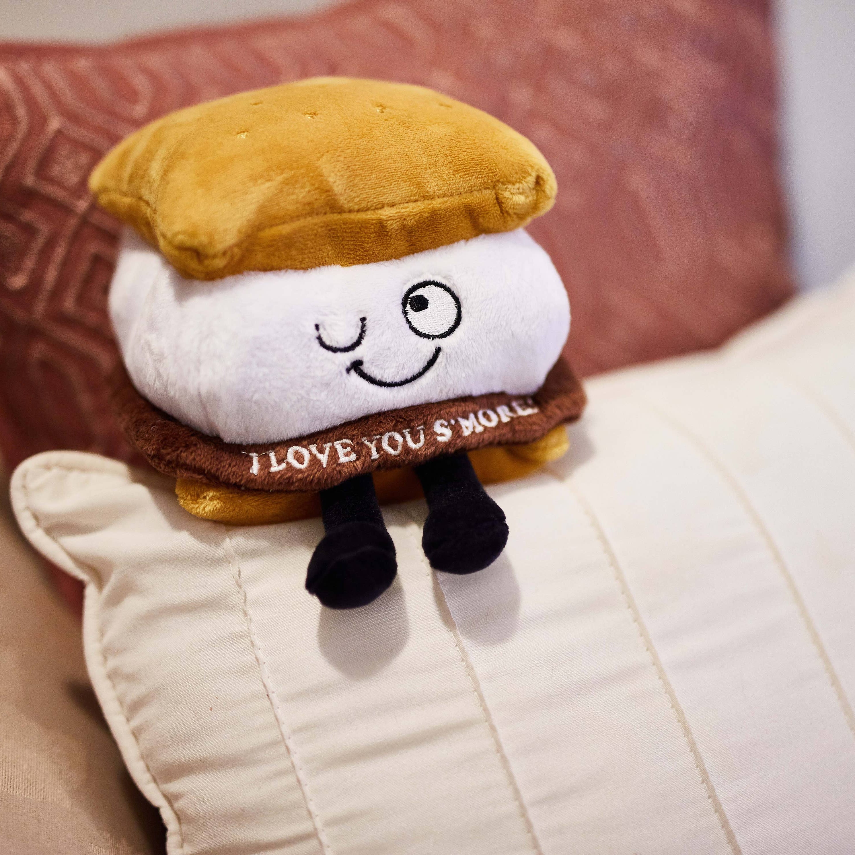 A plushie shaped like a s'more with a winky face. The chocolate layer features white lettering that reads "I love you s'more!"