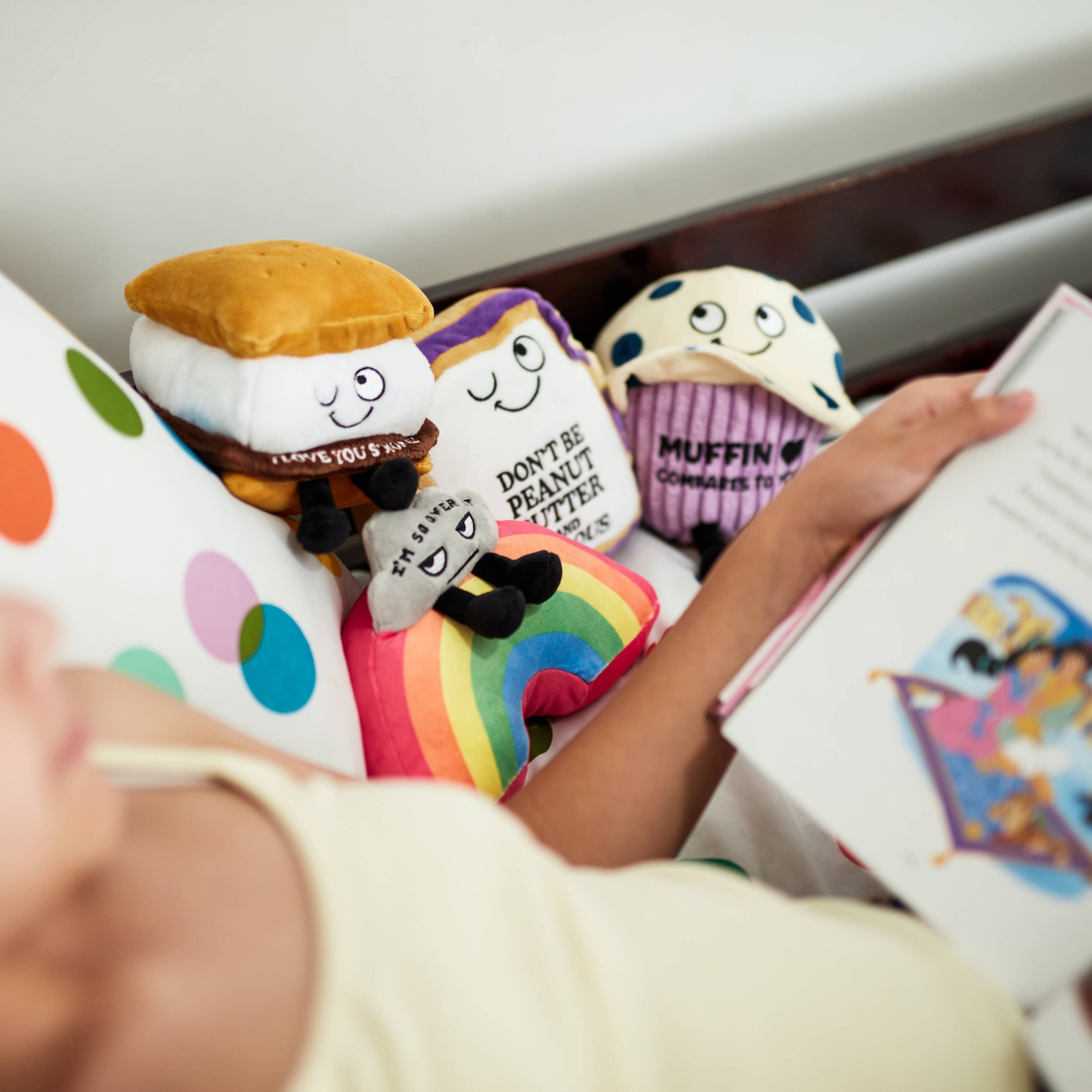A plushie shaped like a s'more with a winky face. The chocolate layer features white lettering that reads "I love you s'more!" Pllushie is sitting on a bed with a child reading a book with other plushies.