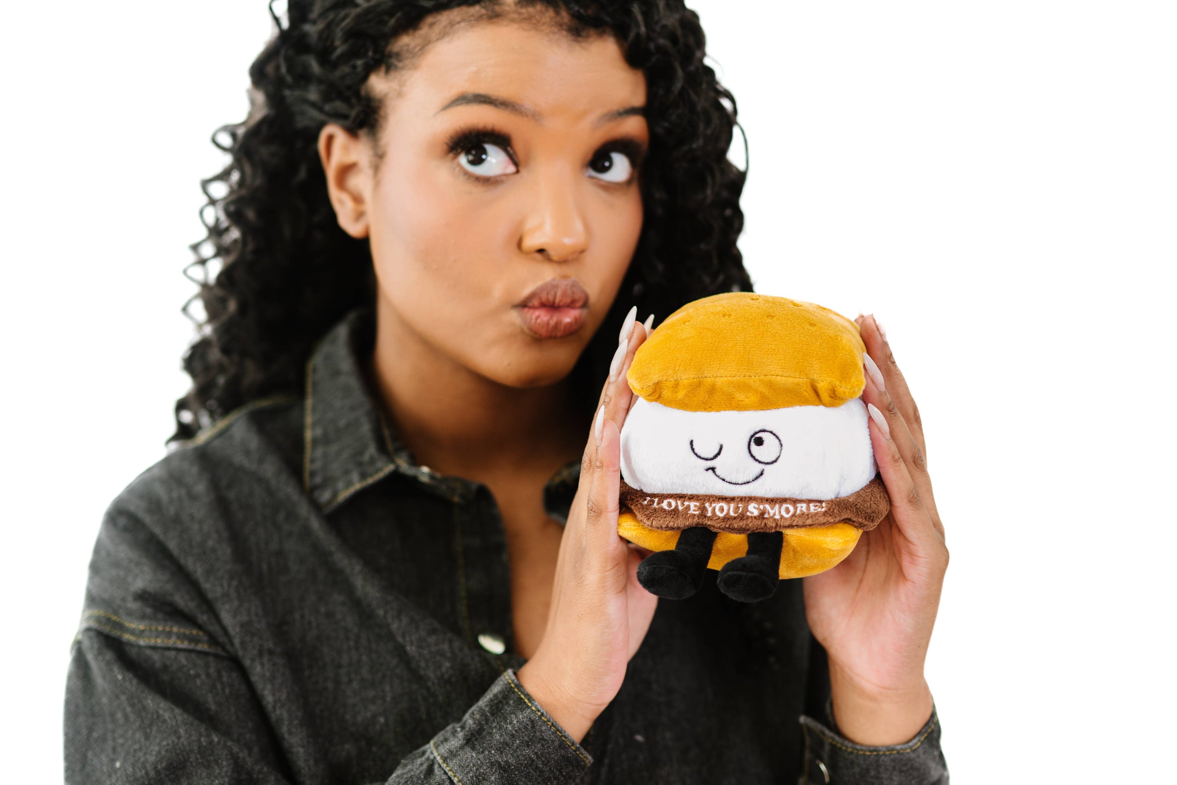 Person holding A plushie shaped like a s'more with a winky face. The chocolate layer features white lettering that reads "I love you s'more!"