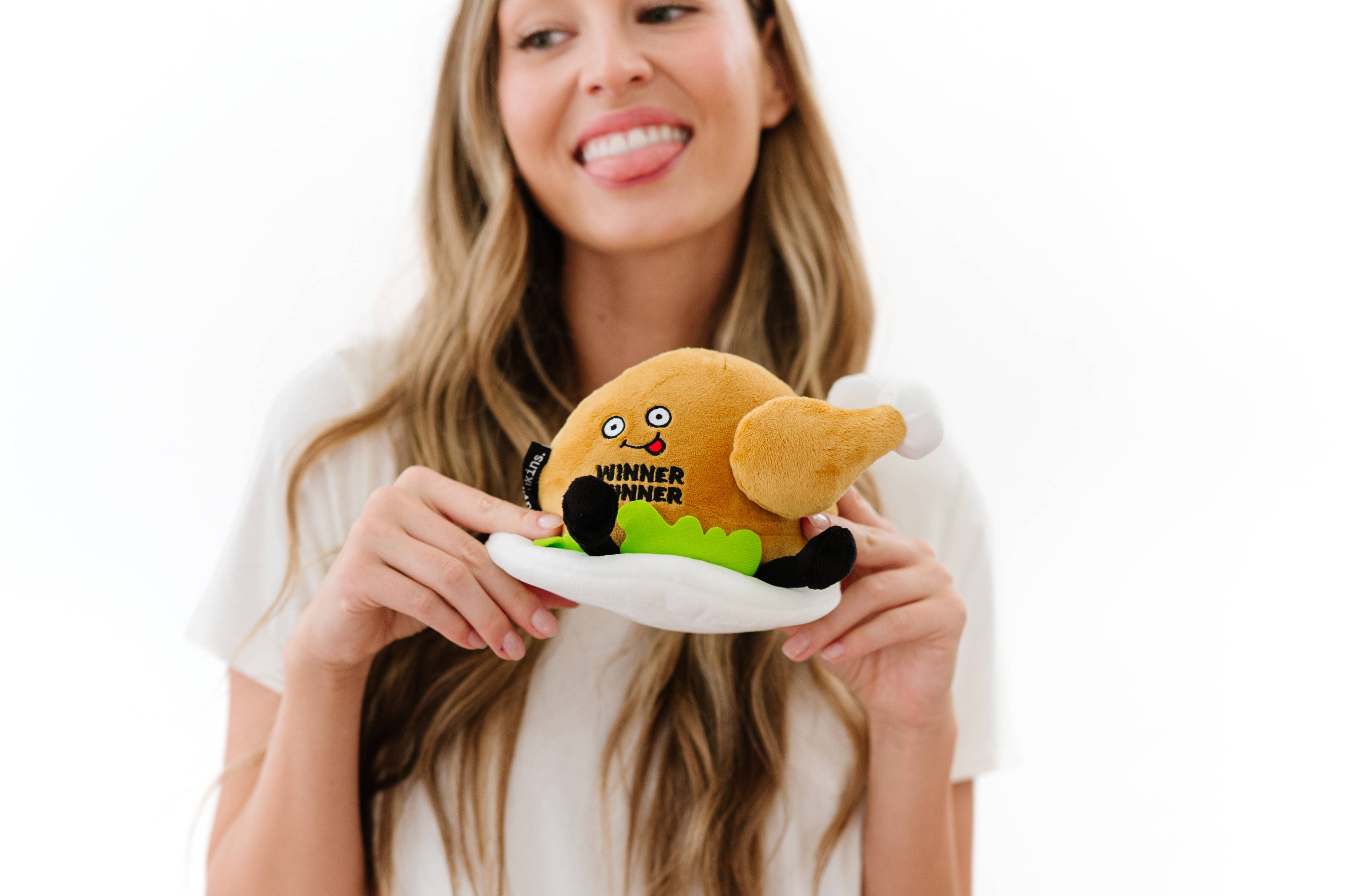 Woman sticking her tongue out while A cooked turkey dinner plush on a plate with green lettuce. The turkey has a smiley face with a red tongue sticking out and black text that says "Winner Winner." It also has black legs sticking out.