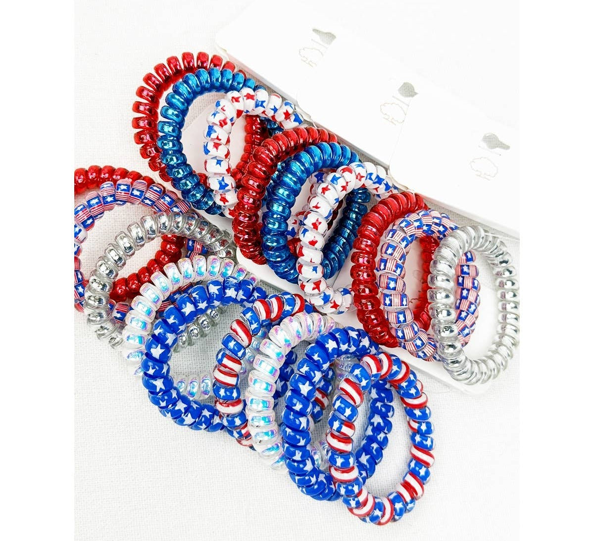 A multi-pack display of spiral phone cord hair ties featuring patriotic United States themes, including solid red, solid blue, white with red and blue star patterns, and silver glitter coils.