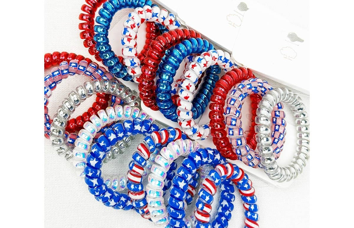 A multi-pack display of spiral phone cord hair ties featuring patriotic United States themes, including solid red, solid blue, white with red and blue star patterns, and silver glitter coils.