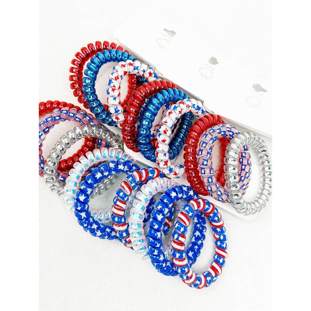 A multi-pack display of spiral phone cord hair ties featuring patriotic United States themes, including solid red, solid blue, white with red and blue star patterns, and silver glitter coils.