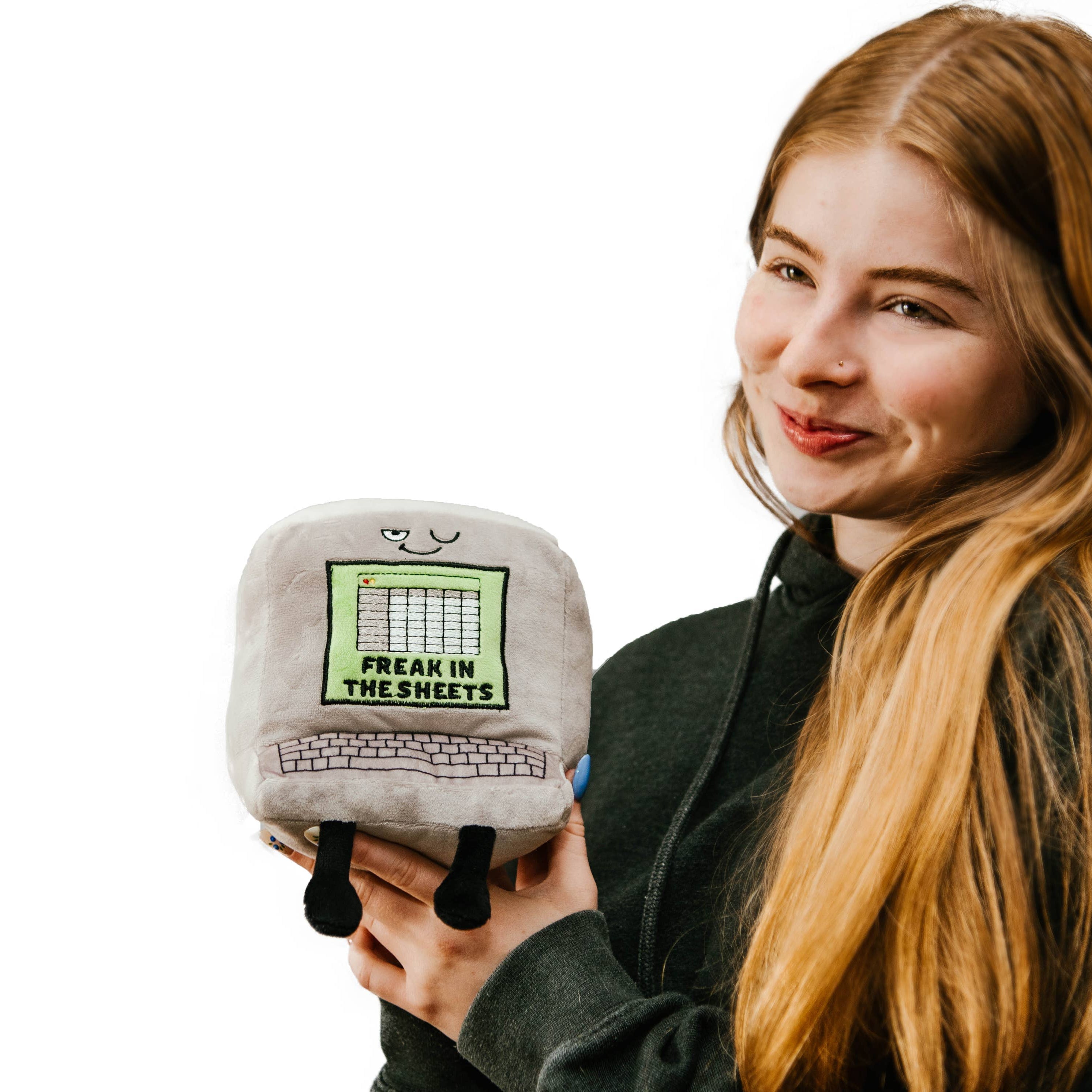 Woman holding a gray computer plush with a winky face, featuring a green Excel-style sheet embroidered with the words "Freak in the Sheets" in black. It also has an outlined keyboard design and black legs sticking out from the bottom.