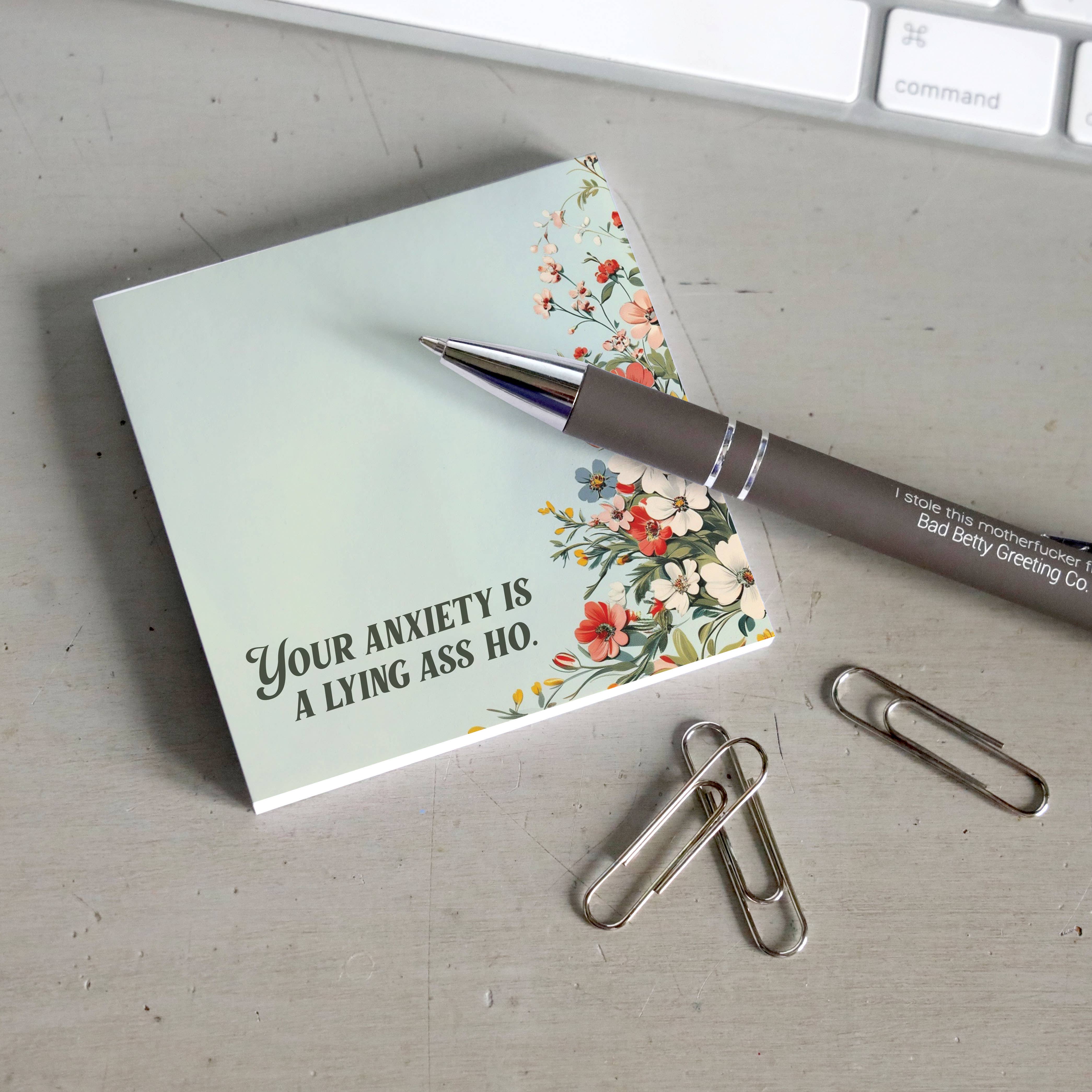 Notebook with a floral design and humorous text on a desk with a pen and paper clips.