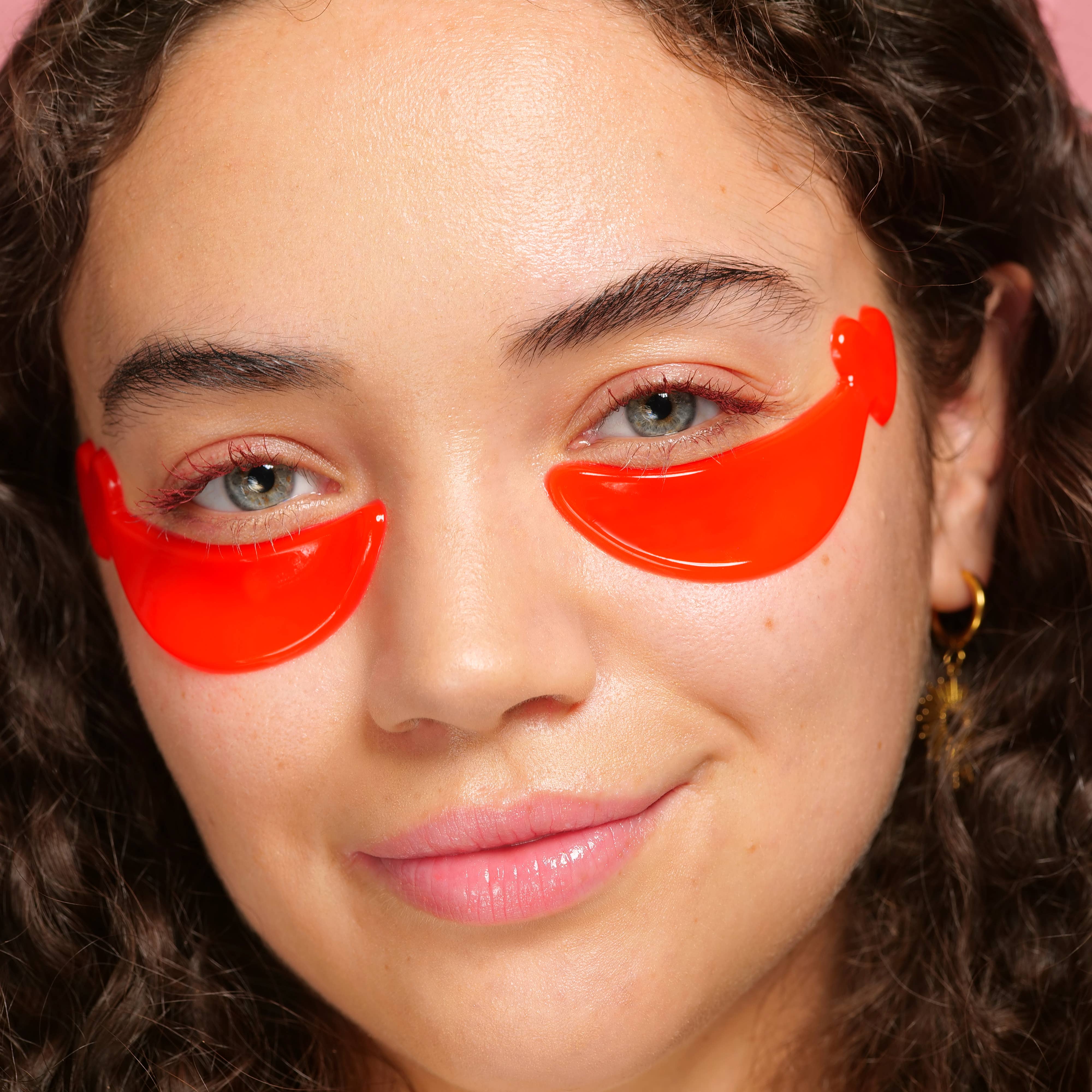 Young woman with curly hair smiles softly, wearing bright red under-eye patches shaped like lips against a neutral background. Her skin looks fresh.