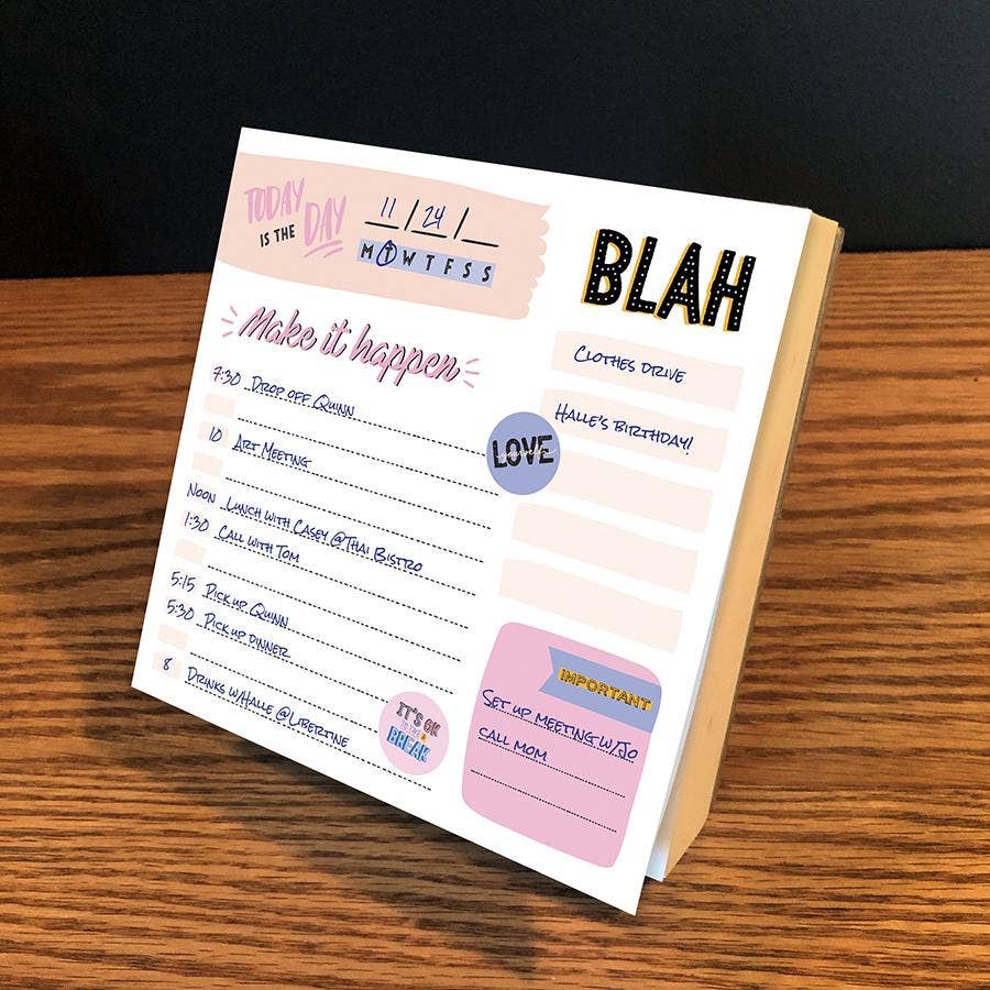 A colorful desk planner page on a wooden surface displays a daily schedule with handwritten tasks. Words like "Blah" and "Love" add a playful tone.