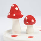 Two ceramic mushrooms with glossy red caps and white spots stand on a white base. The whimsical tones add a playful, decorative touch.