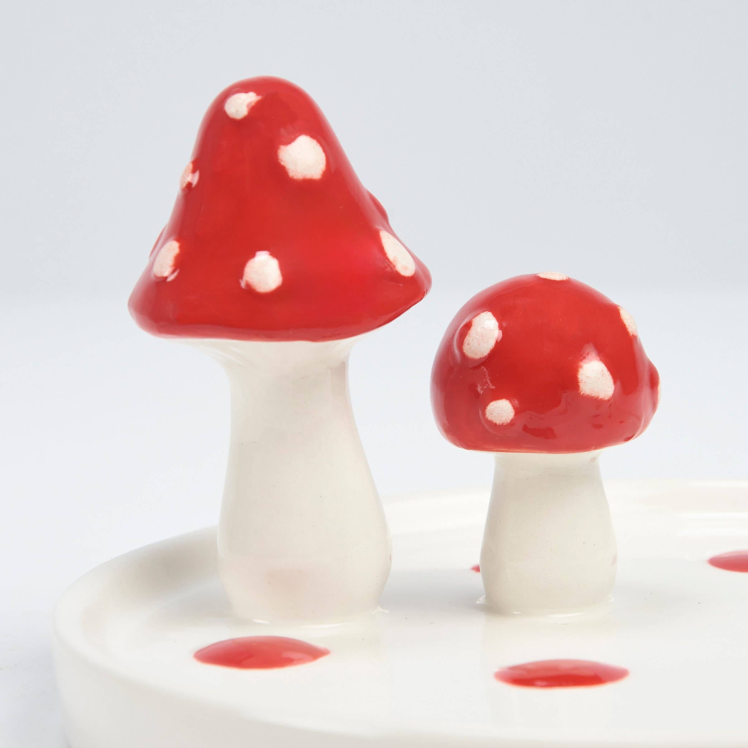 Two ceramic mushrooms with glossy red caps and white spots stand on a white base. The whimsical tones add a playful, decorative touch.