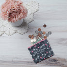 A patterned pouch with the text "I'm not needy, I'm wanty" spills coins and bills on a light wooden table. Nearby, pink flowers sit in a vase on a lace mat.